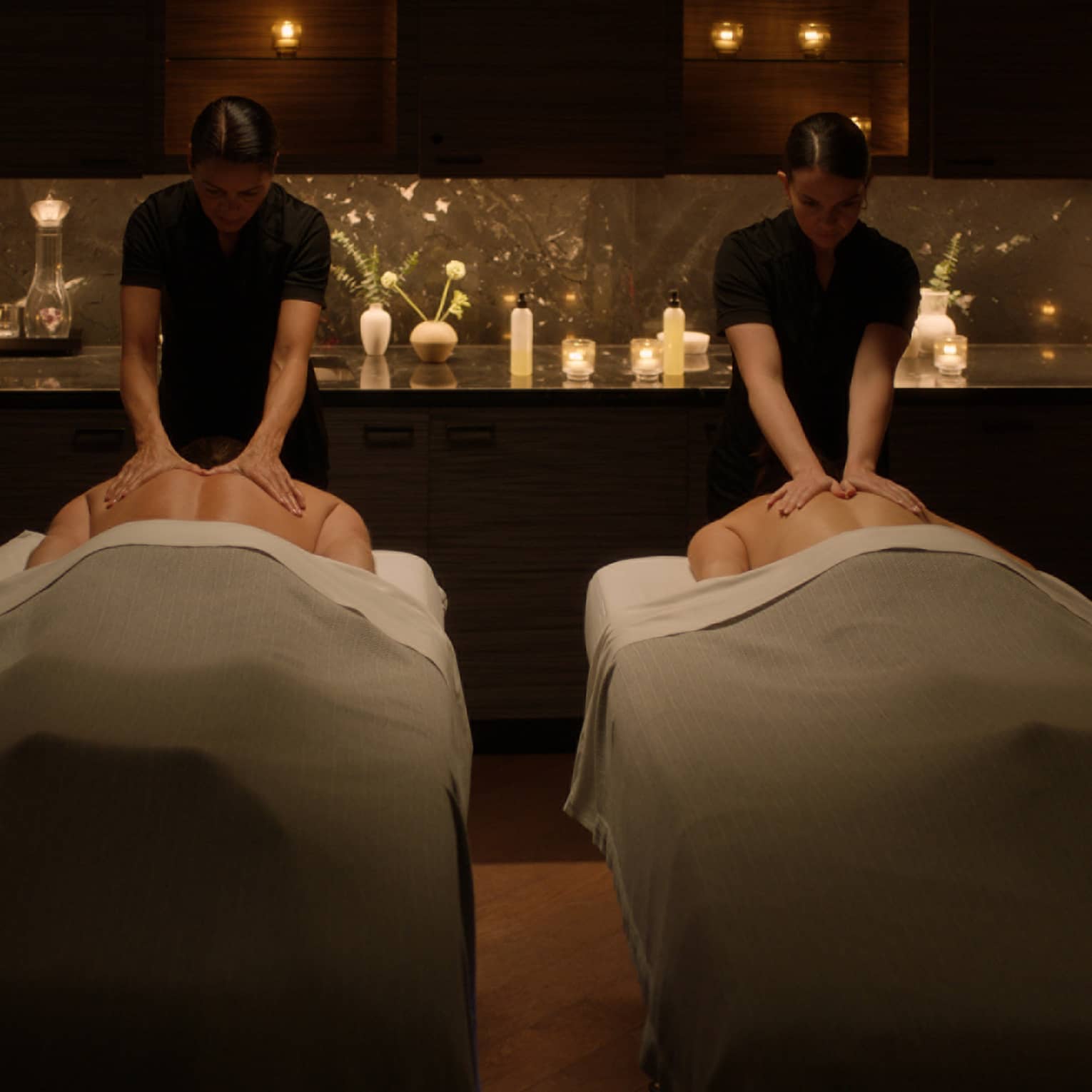 Two people lying face down on massage tables and two massage therapists providing massage in a serene space with low lighting and candles
