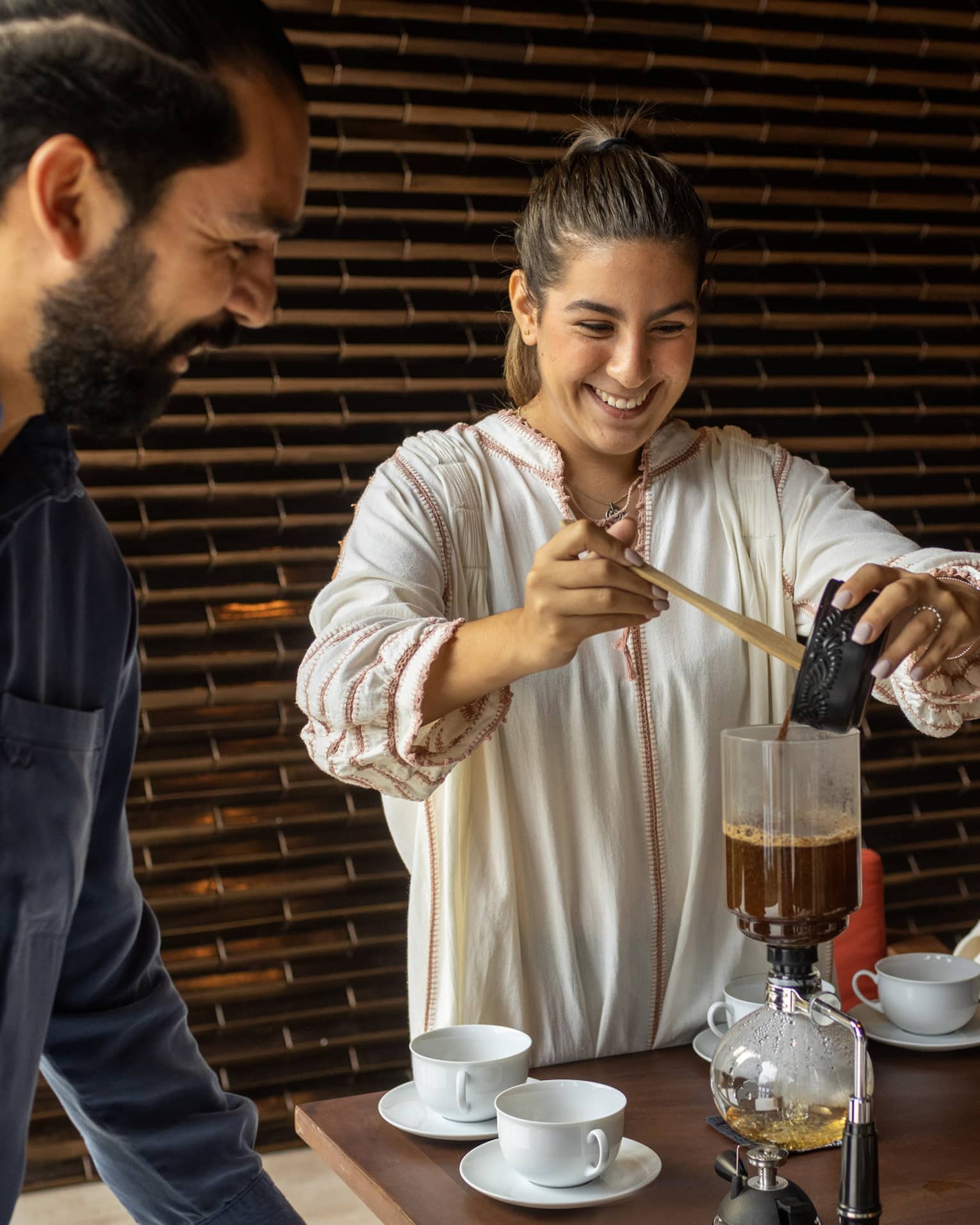 At a table set with white cups and saucers, two smiling guests watch as another pours coffee into a siphon coffee maker.