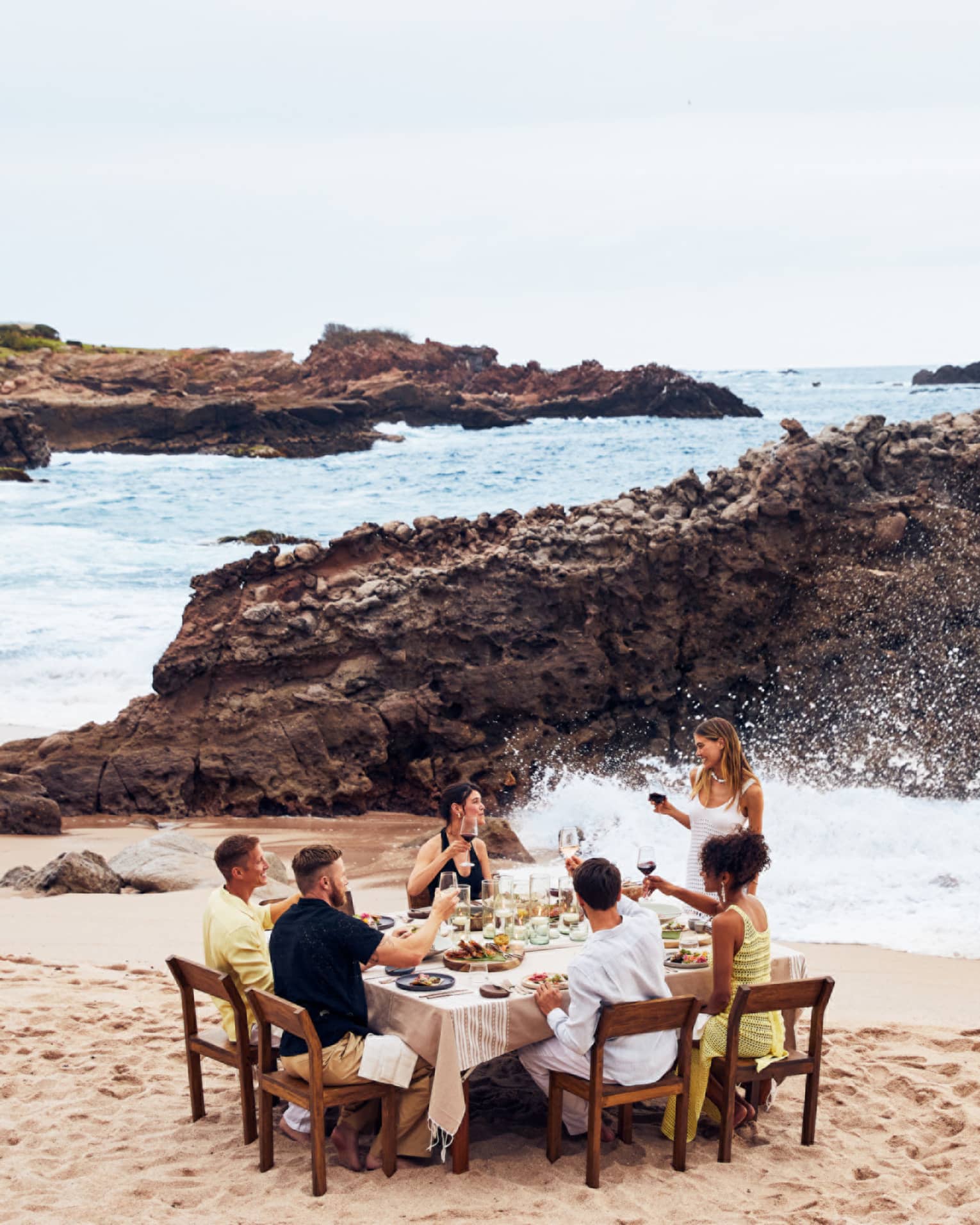 A group eating at a table on a beach with waves crashing nearby.