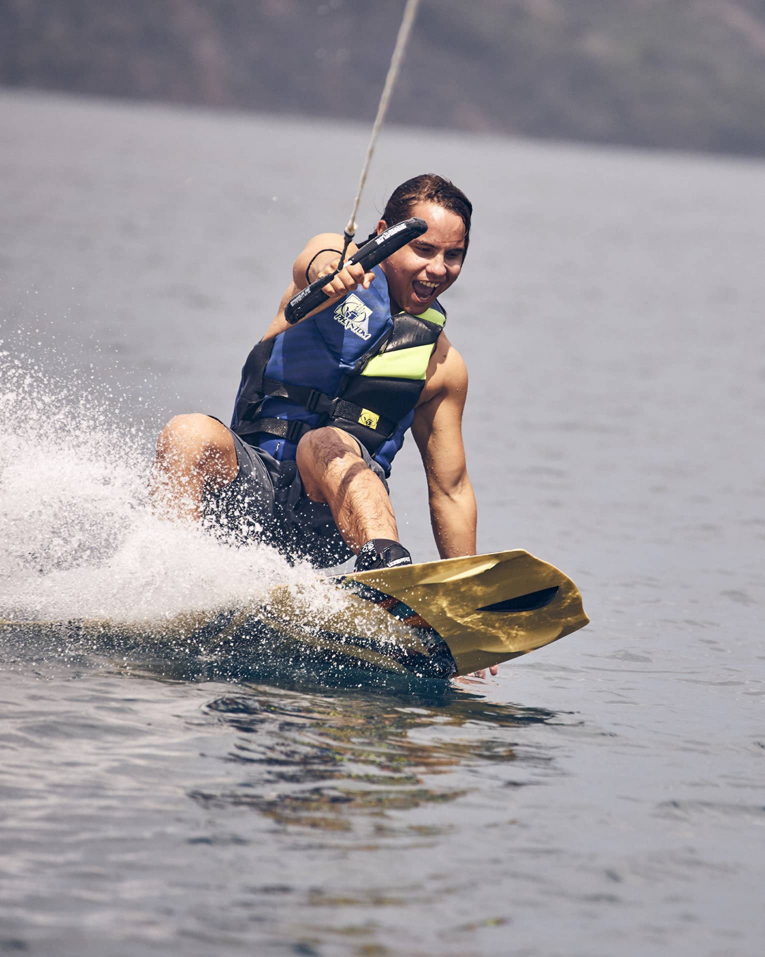 Young man wearing a blue and green life vest and blue swim trunks wakeboarding through the water