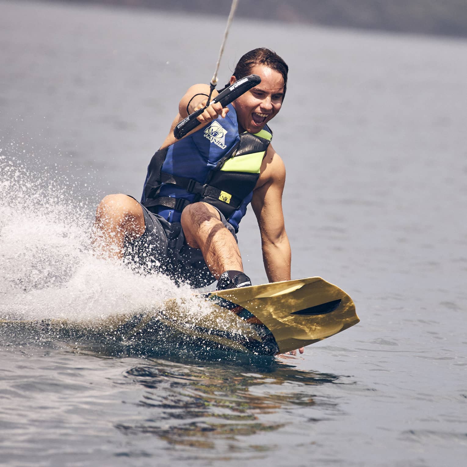 Young man wearing a blue and green life vest and blue swim trunks wakeboarding through the water