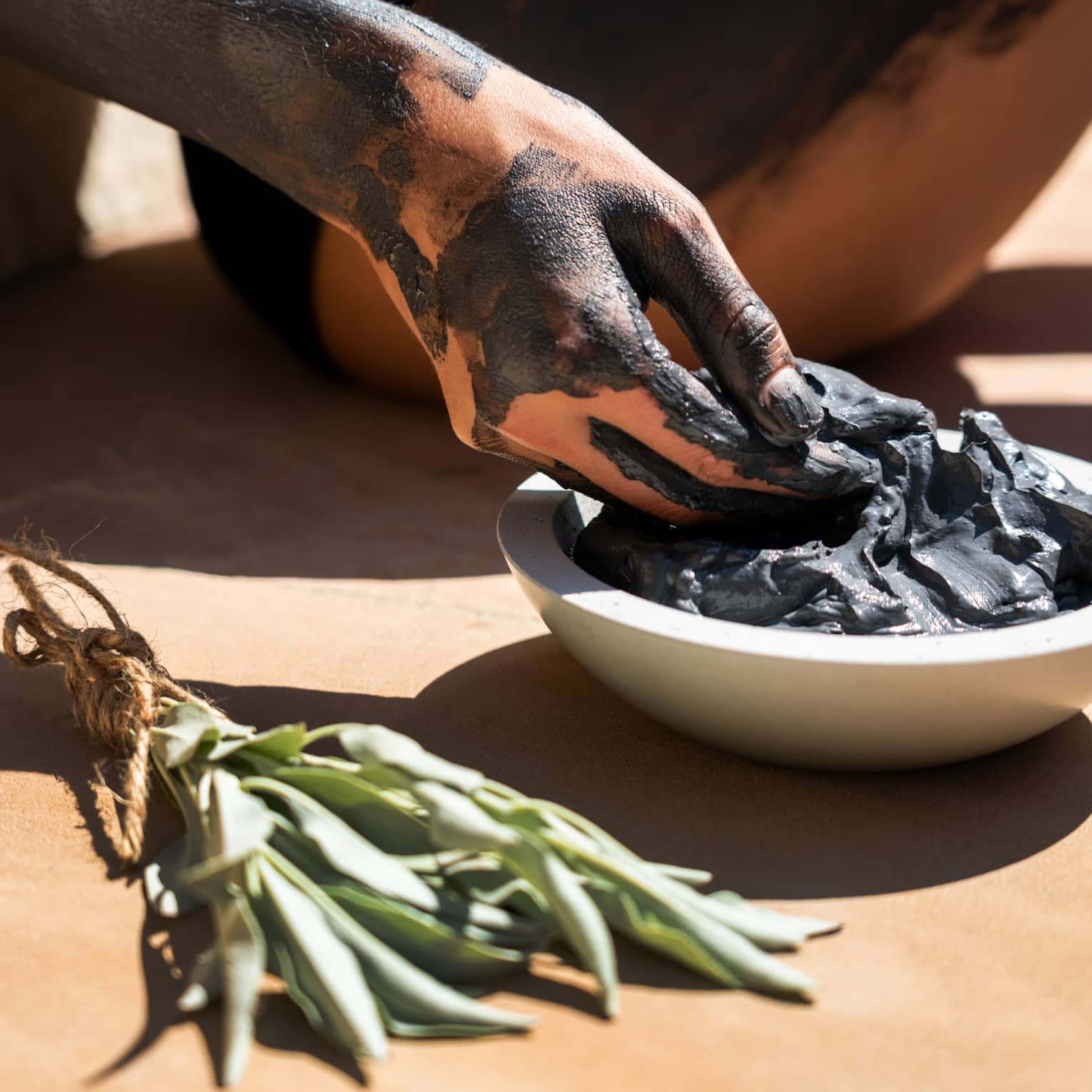 Hand covered in black clay reaching into a bowl of spa mud next to sage bundle