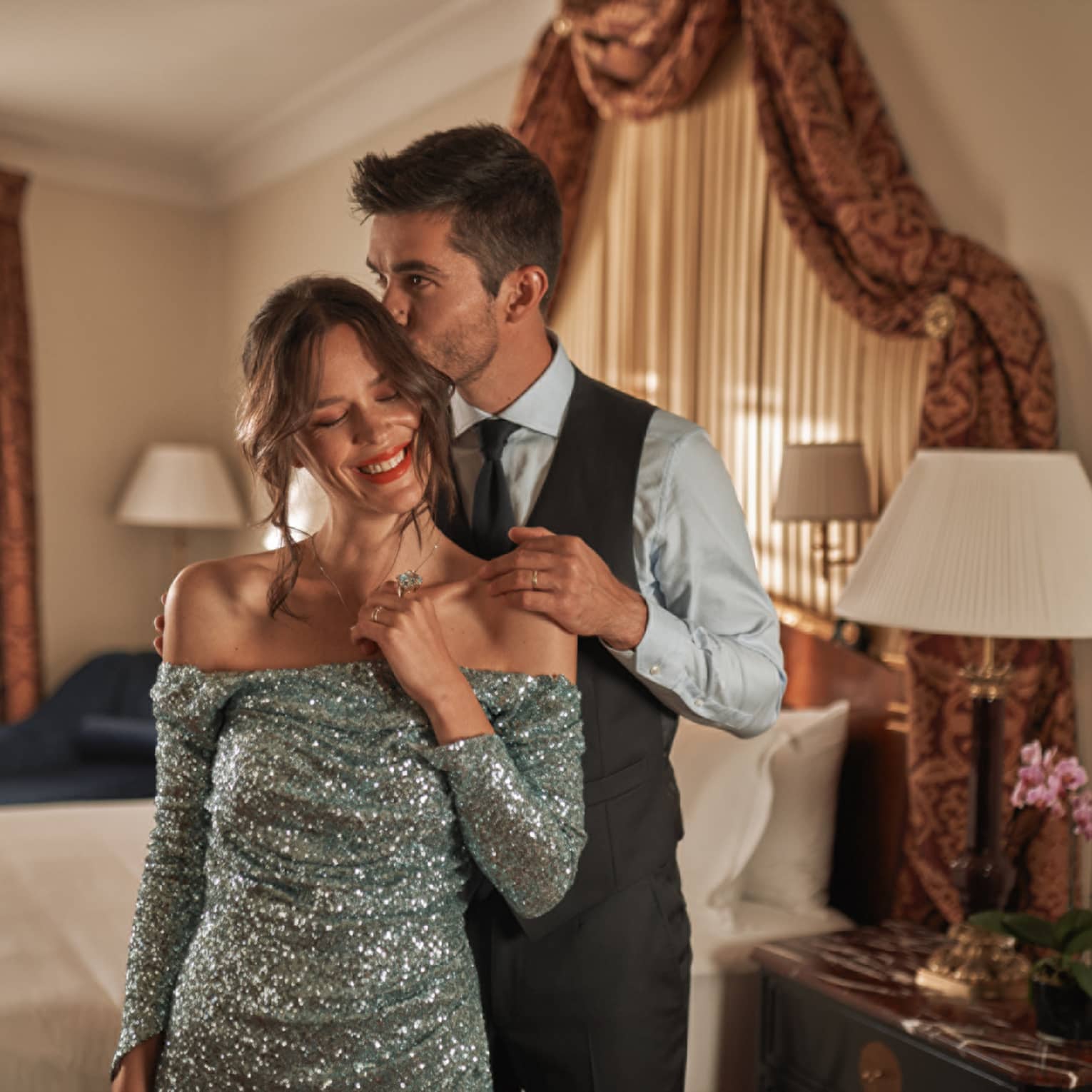 Man in suit and woman in sequin dress stand together in an elegant bedroom