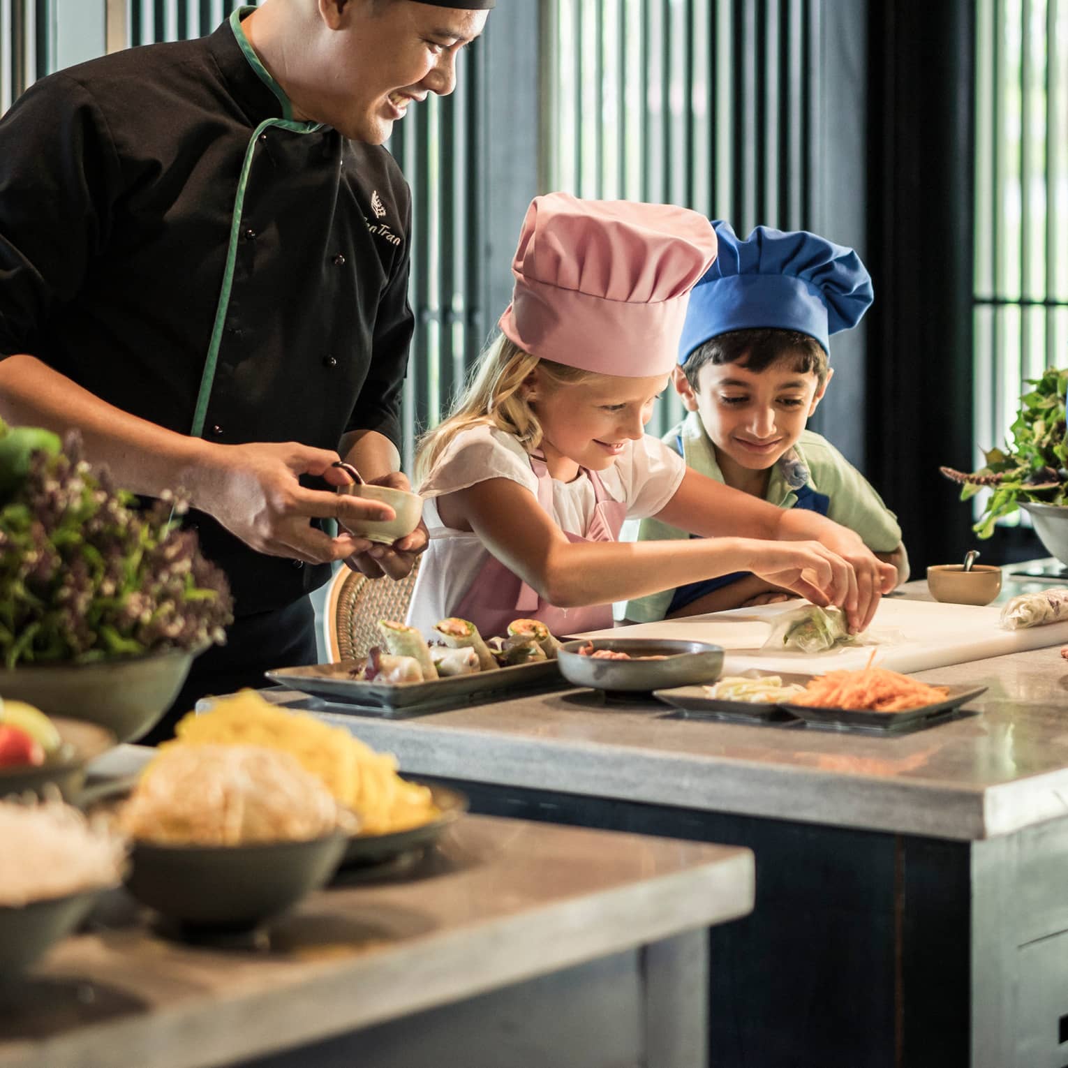 Chef watches as children wearing tall chef hats make spring rolls at counter