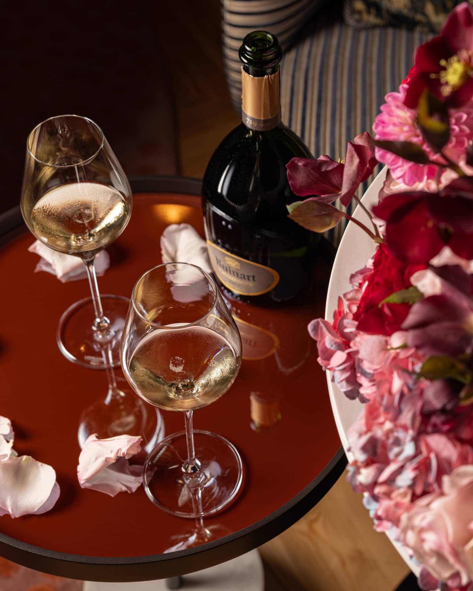 Two glasses of white wine on a brown table, surrounded by scattered rose petals and placed next to a large bouquet of flowers.