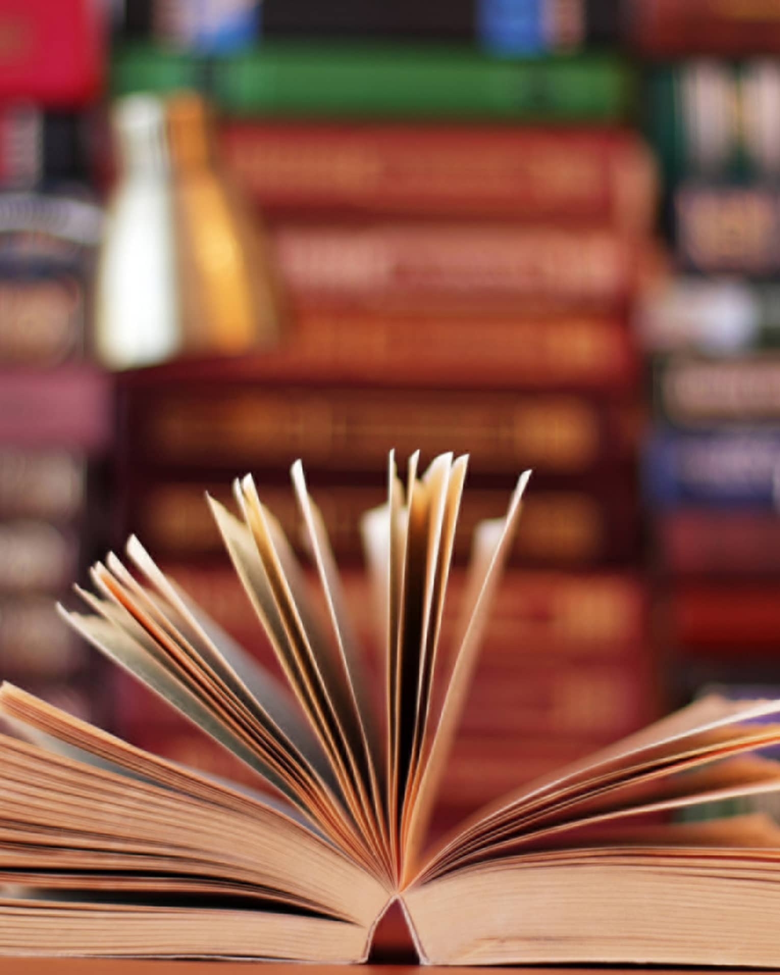 A book is opened, with pages fanned, lamp and stacks of books in background 