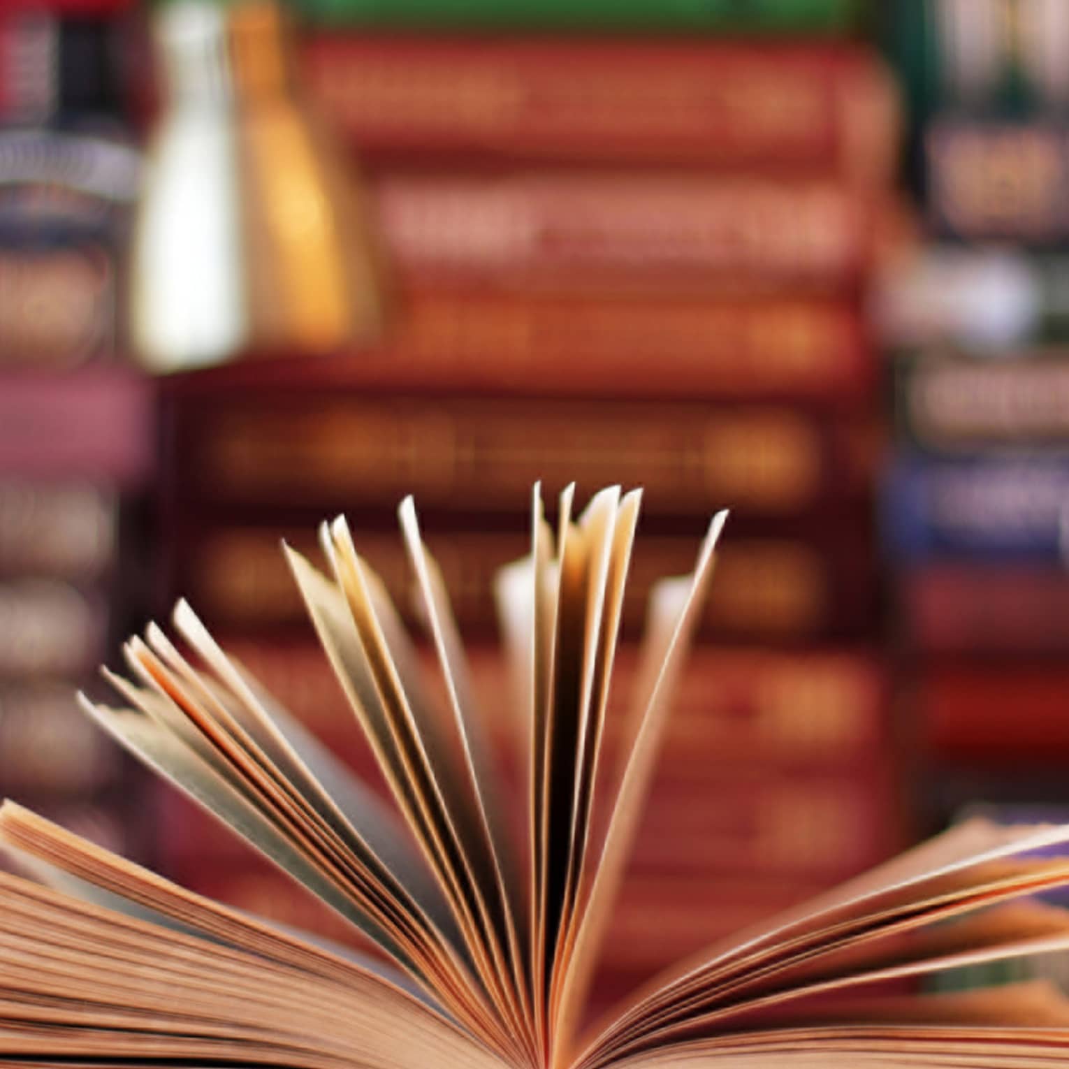 A book is opened, with pages fanned, lamp and stacks of books in background 
