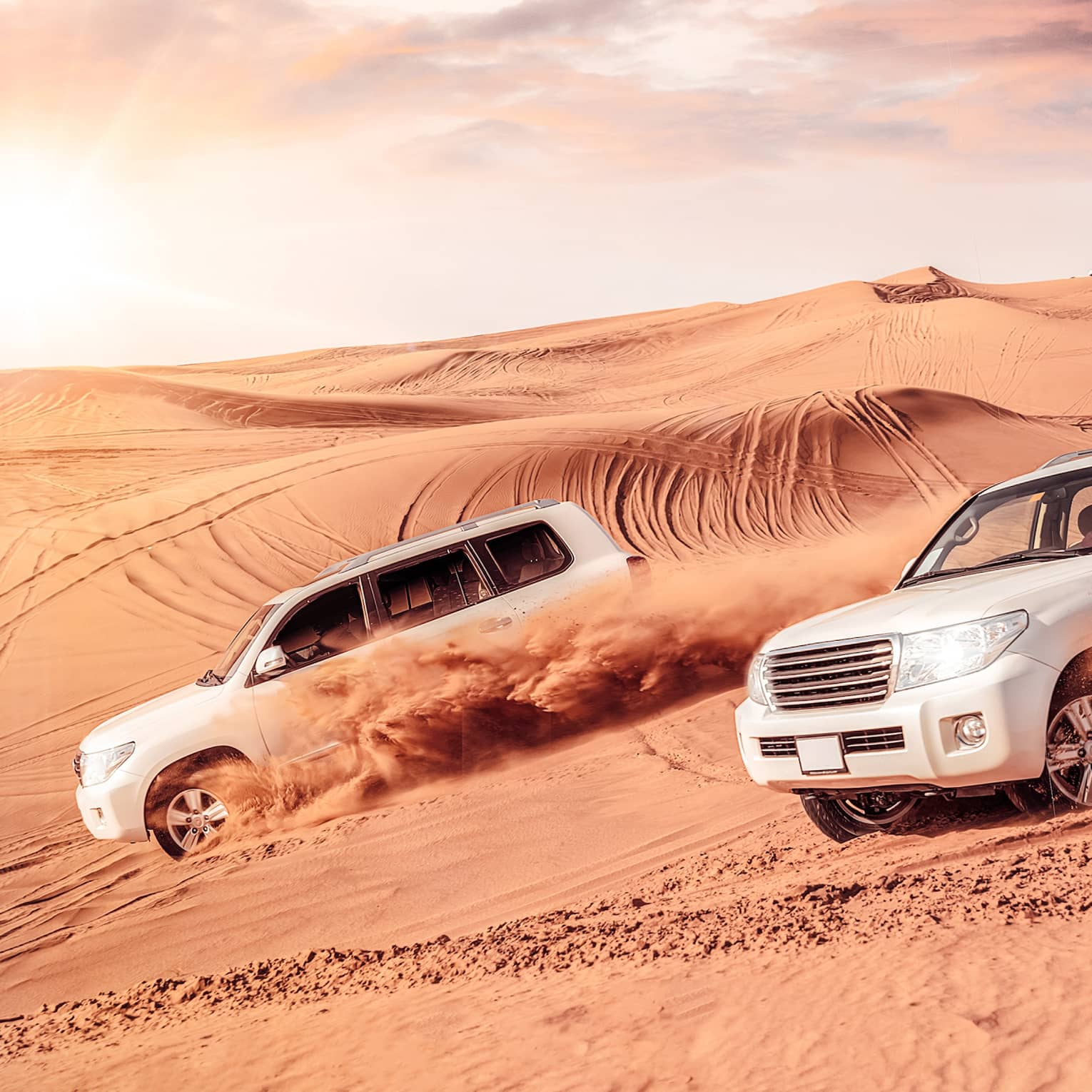 Sand flies from tires of two white SUV's driving across dunes