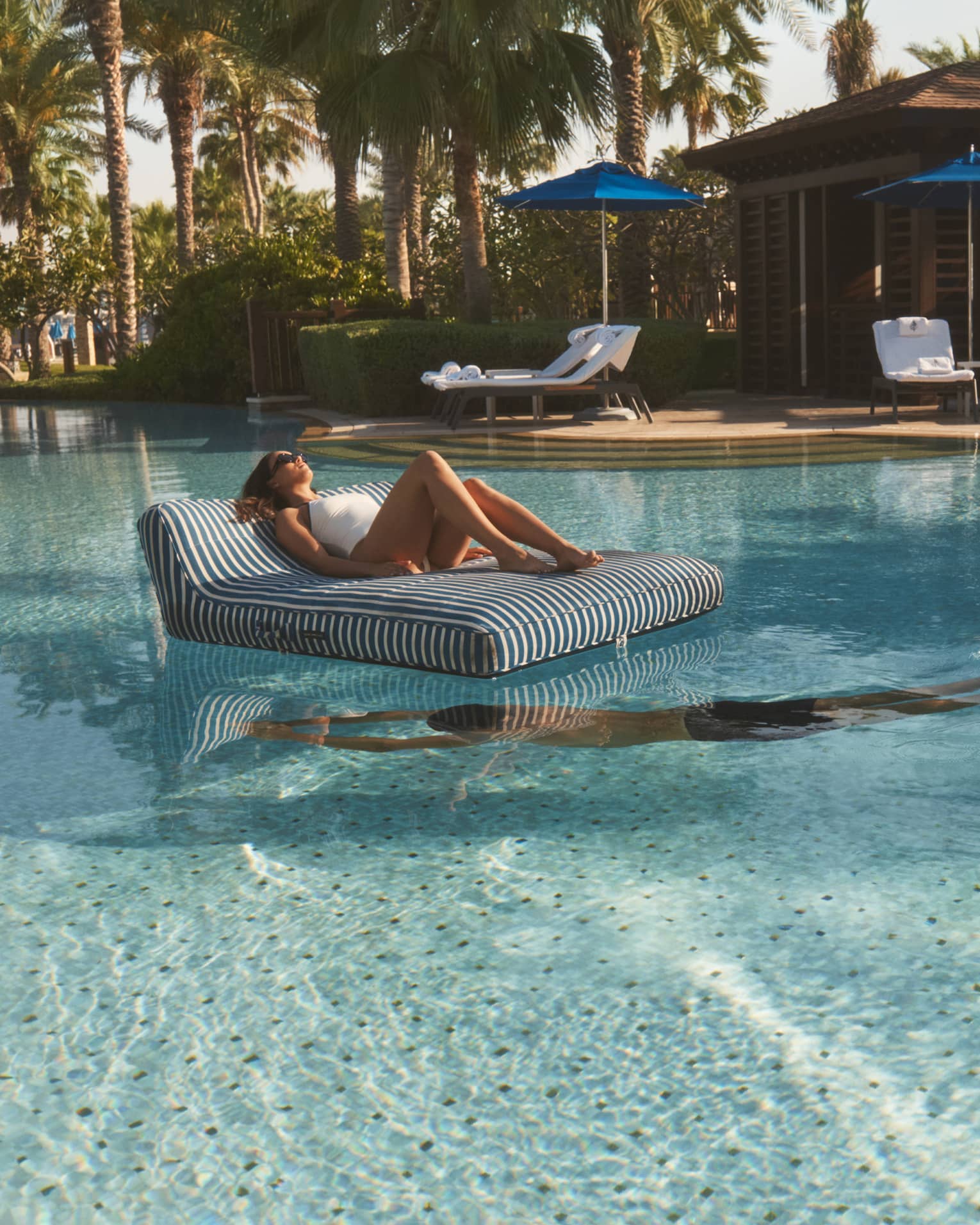 Person lounges on a black-and-white striped floating chair in the middle of a pool as another person swim beneath