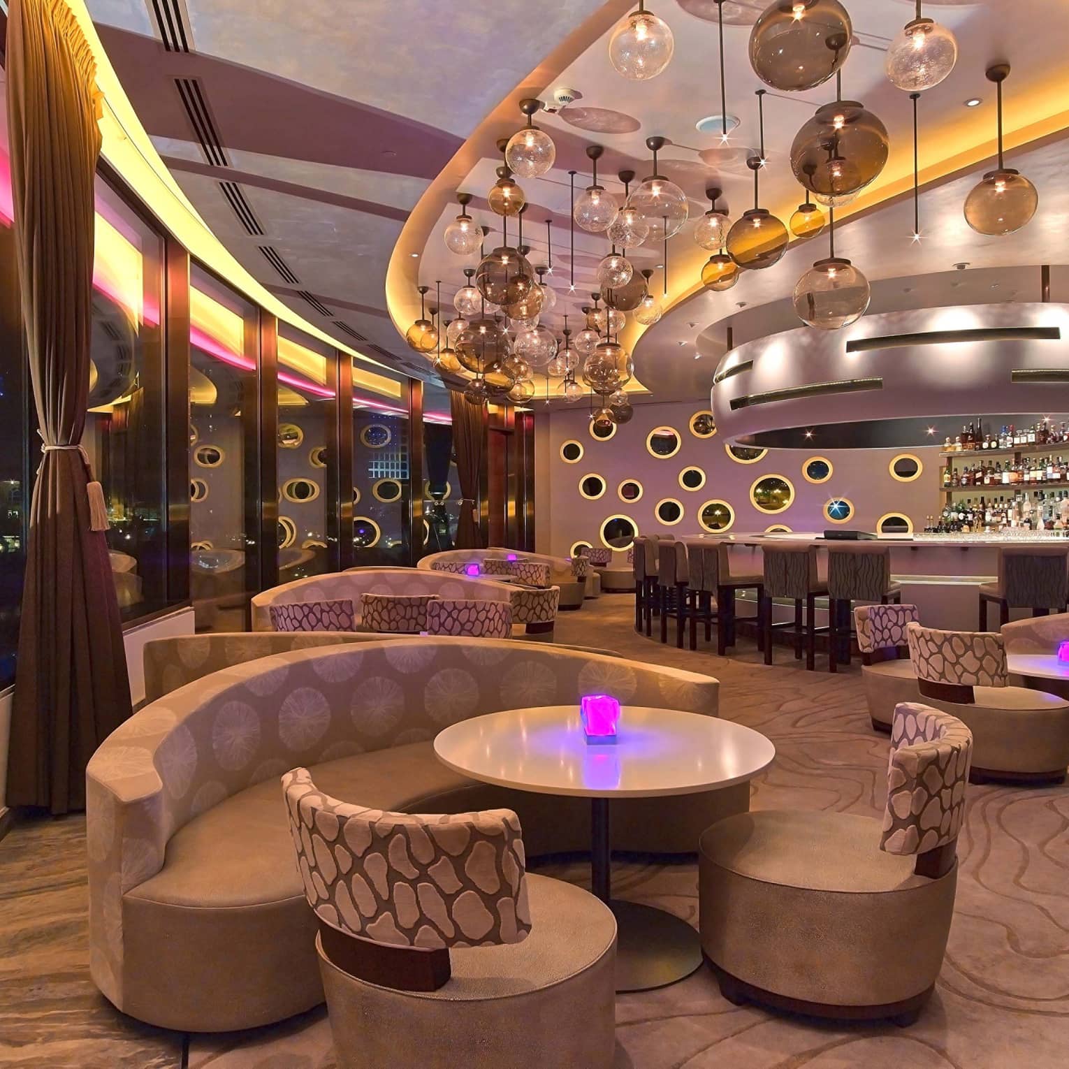 Nighttime interior of Nobu Doha featuring curved seating, cocktail tables, round glass lighting fixtures hanging from the ceiling and pink, purple and yellow lighting accents