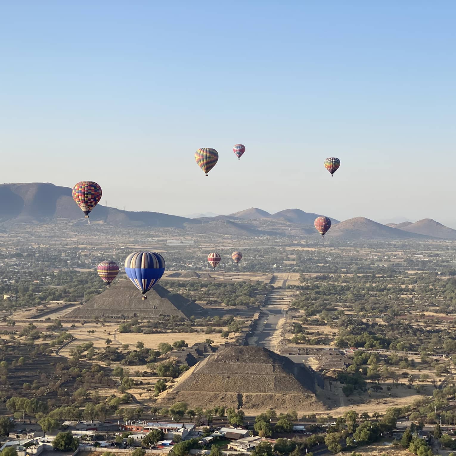Hot-air balloons floating over old temples in a dessert covered in shrubs.