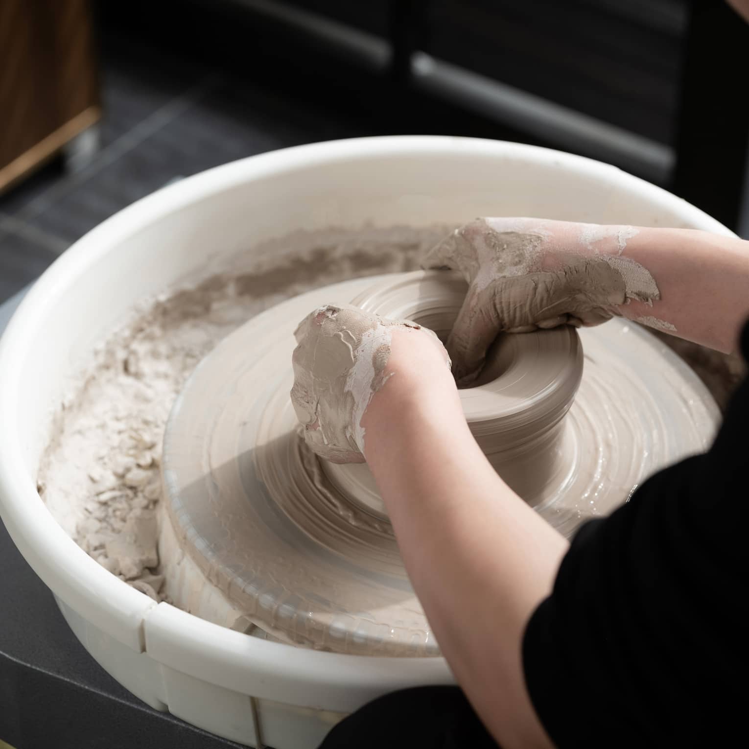 Close-up of two hands covered in wet beige clay shaping a pot on a spinning pottery wheel. The potter is wearing black.