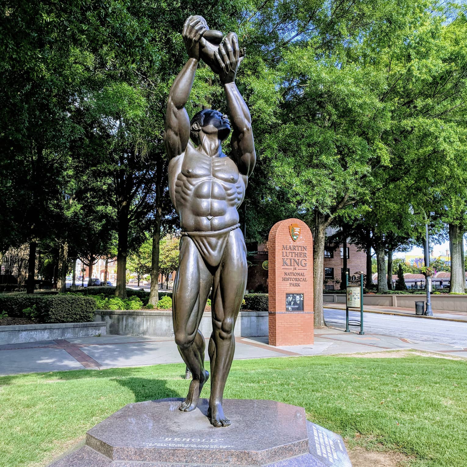 A statue of a man holding something in the air placed in a park with green trees surrounding it.