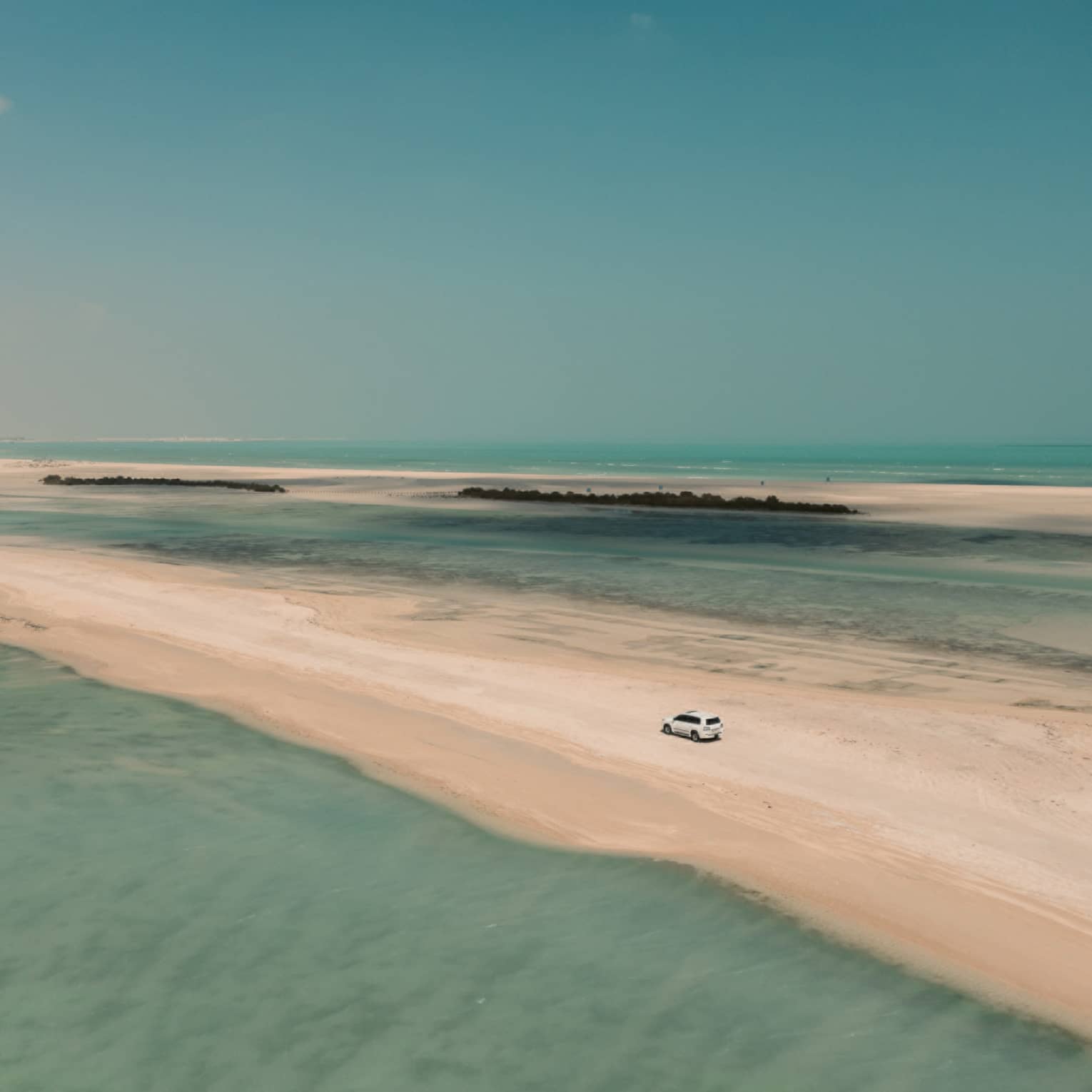 Aerial view of a 4x4 car traversing a sandbar surrounded by shallow turquoise water on either side and a cloudless sky above.