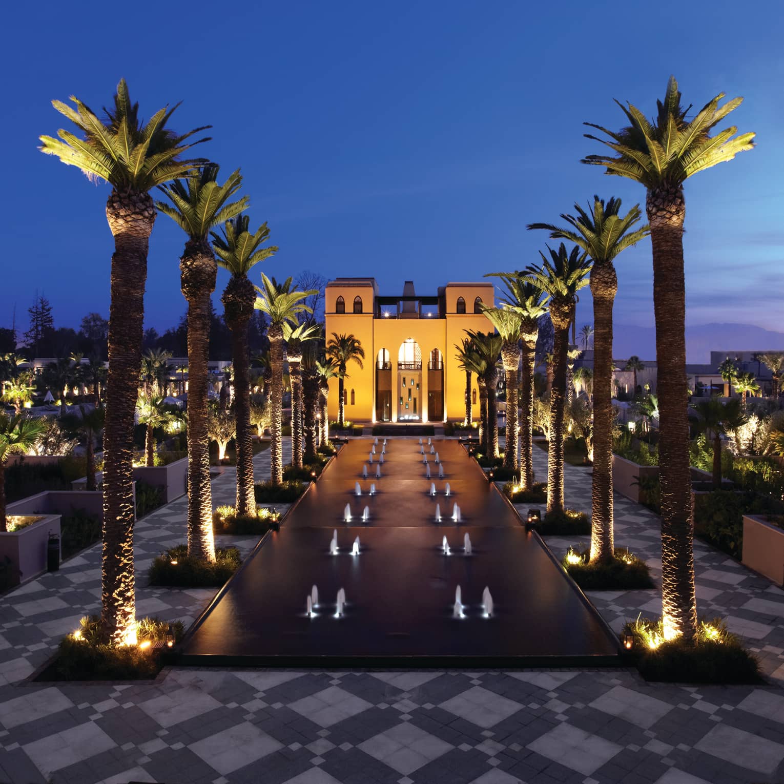 Fountains between rows of tall palm trees in front of illuminated Four Seasons Resort Marrakech hotel at night