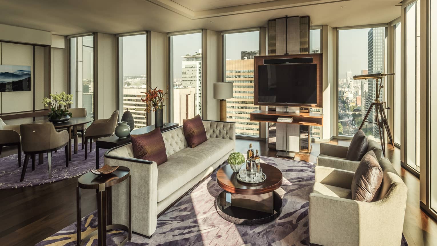 Ambassador One-Bedroom Suite long sofa, brown silk pillows, two armchairs around table with liquor, corner windows and telescope
