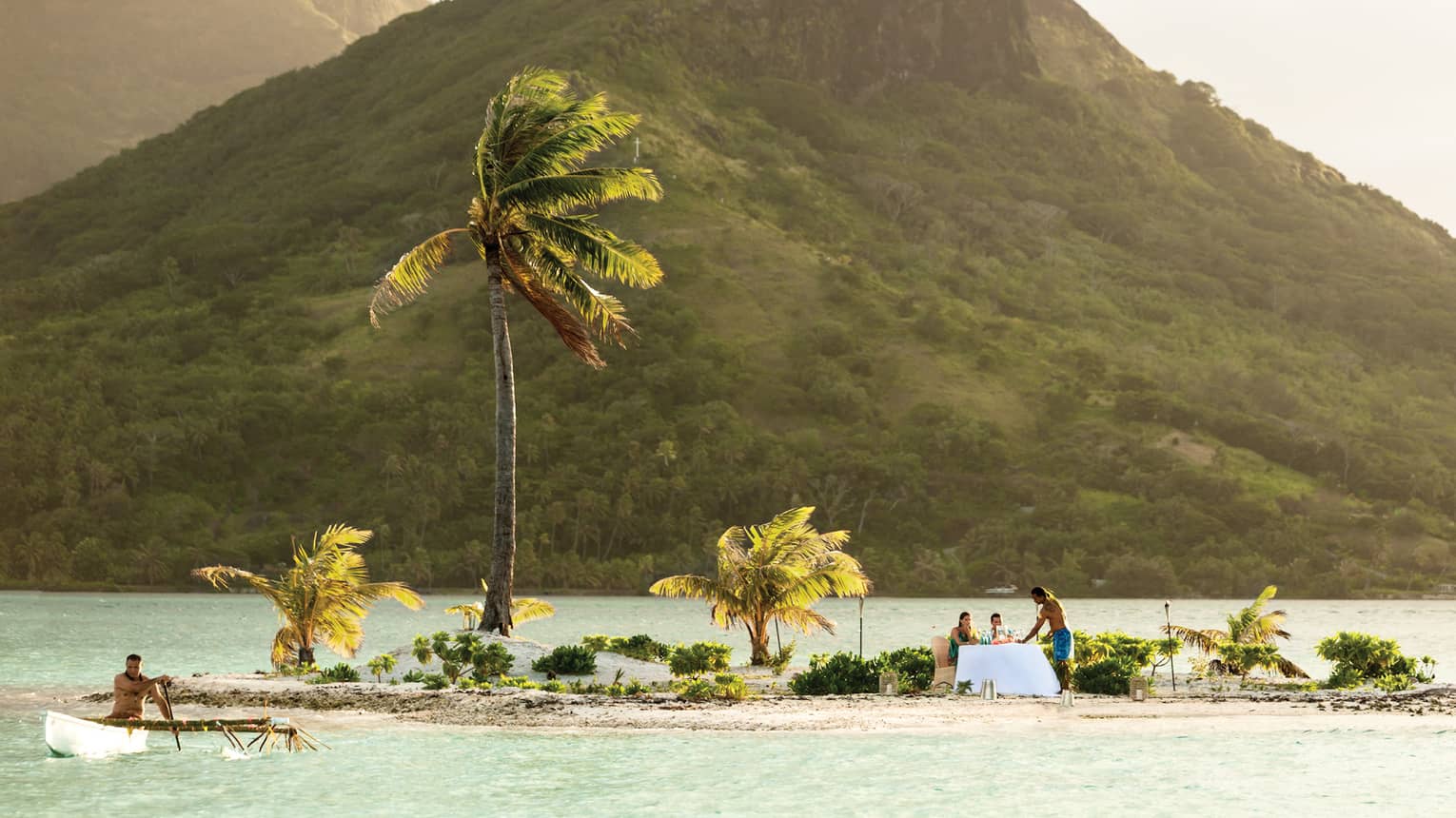 Small tropical island with palm trees, where two people are dining at a table by the water, with a mountain in the background
