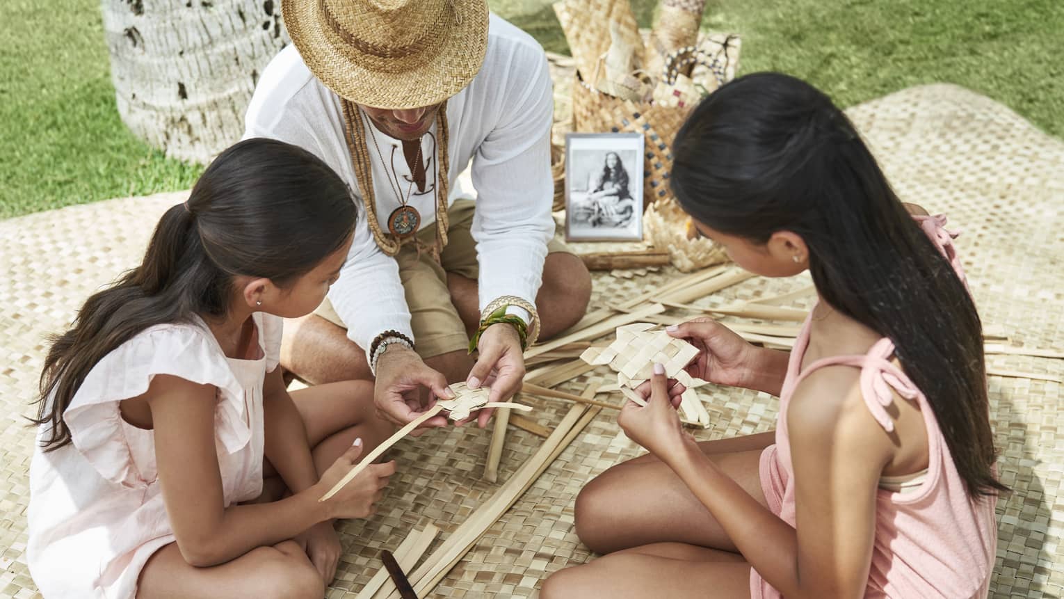 Adult helps two young girls learn how to weave as they sit on a woven blanket