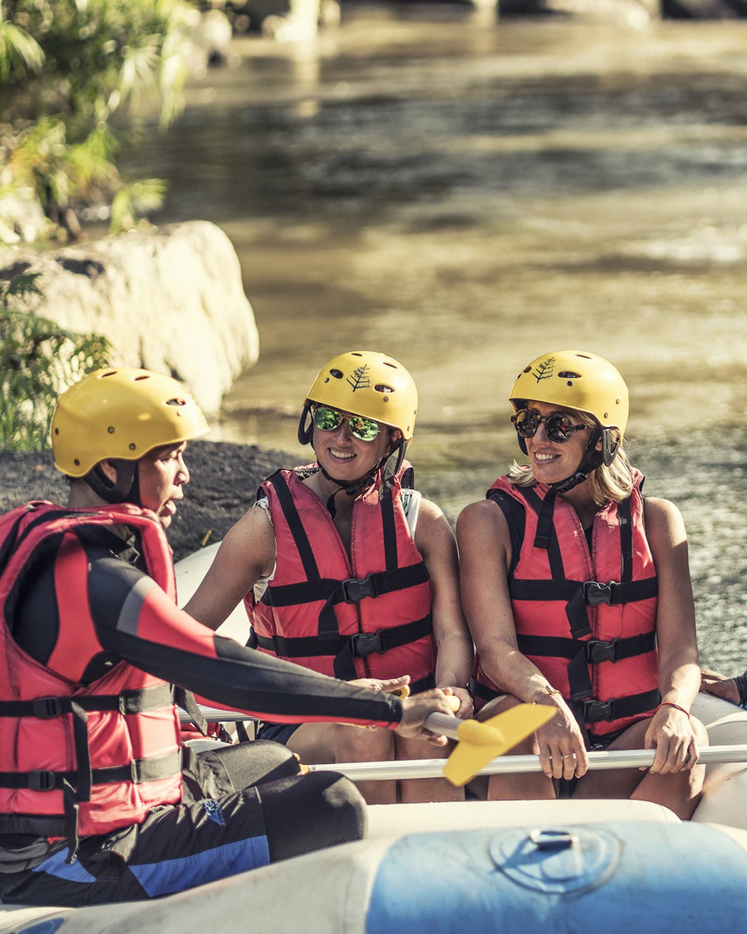Four people wearing red life jackets, sunglasses and yellow helmets sit facing each other in a river raft