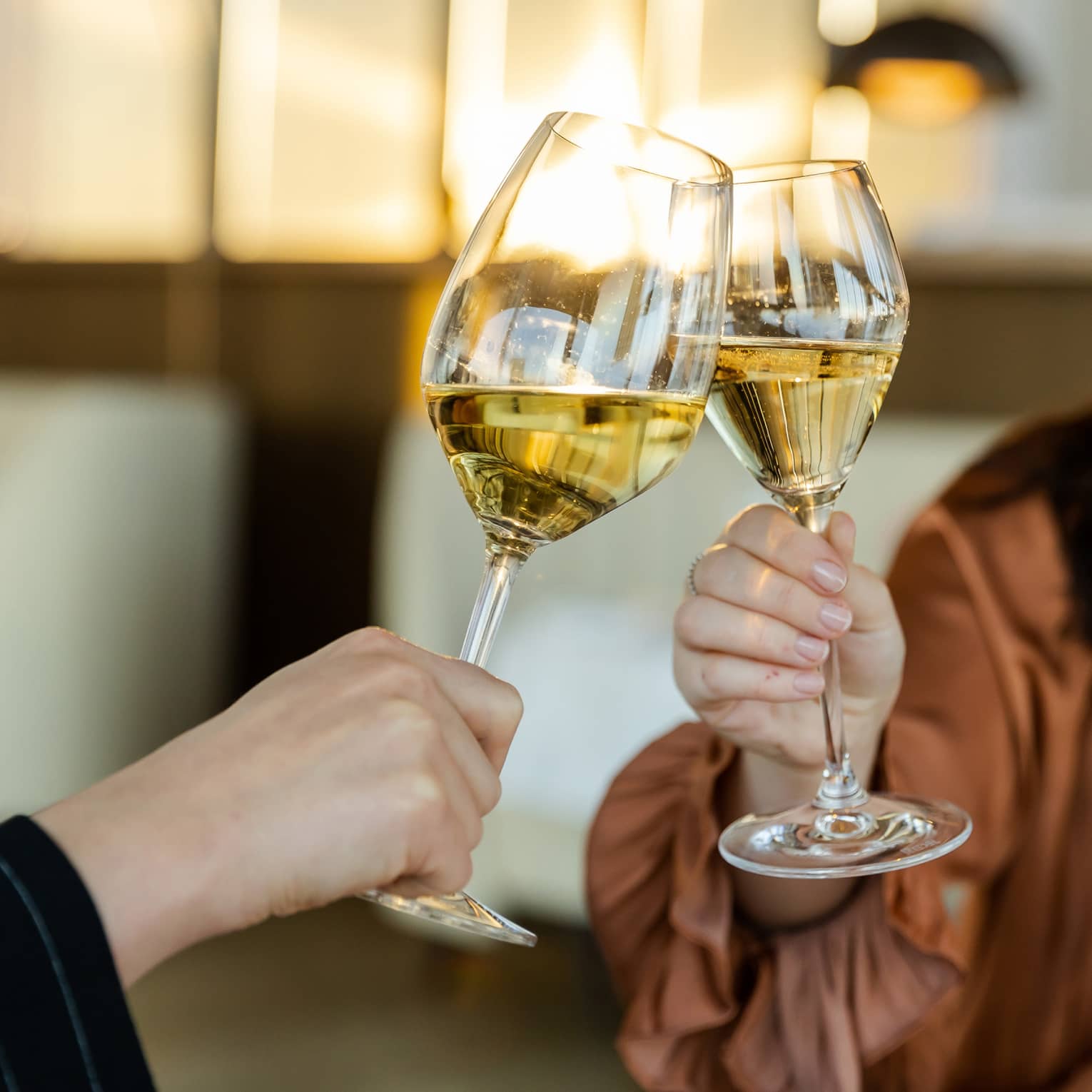 Close-up of two hands clinking glasses of white wine together; beyond, a person in flowing blouse and long brown hair smiles.