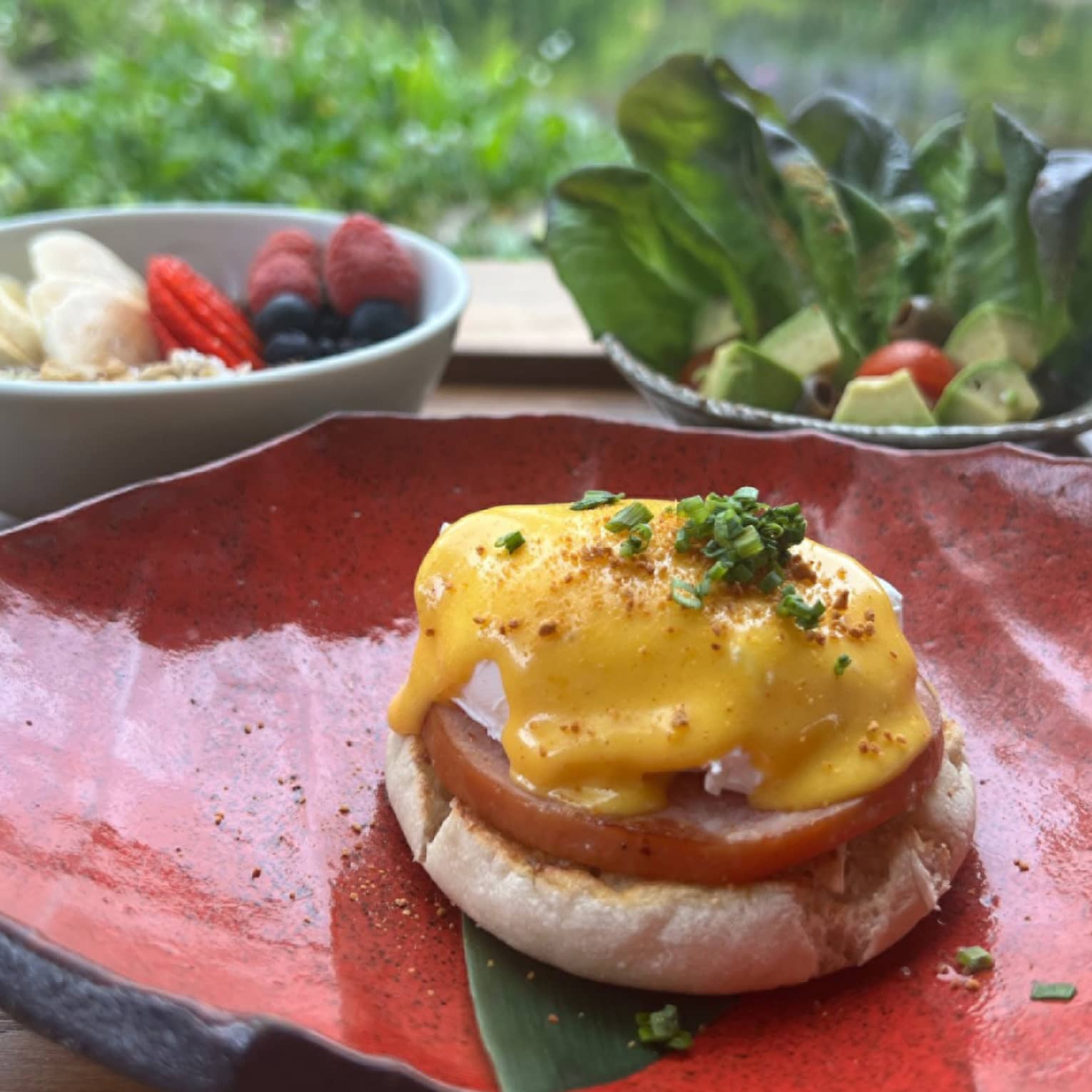 Eggs Benedict on a red stone plate, plus a parfait and salad on a wooden outdoor table