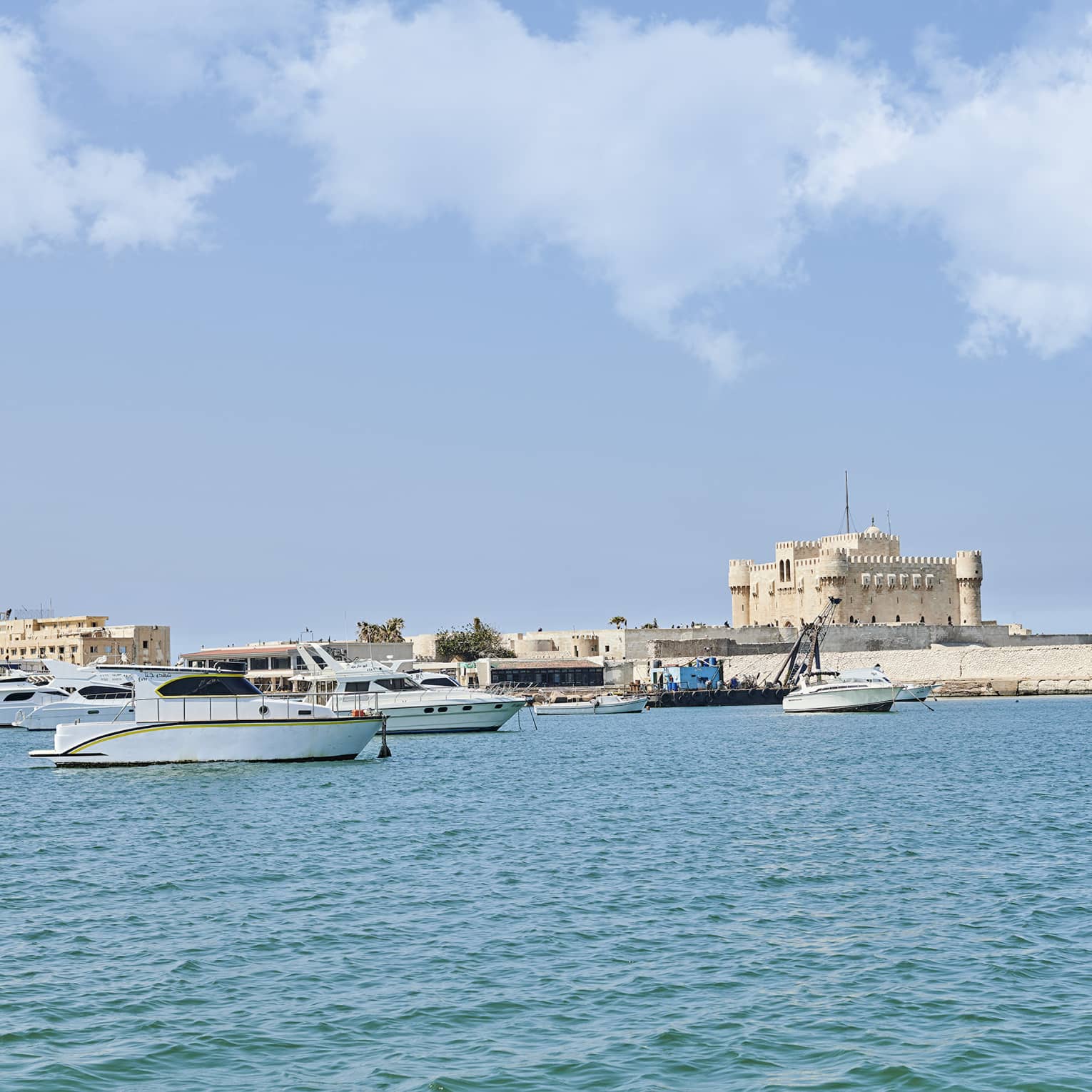 A distant sand-coloured fortress perched on a quay along a shore dotted with small yachts and fishing boats over calm waters.