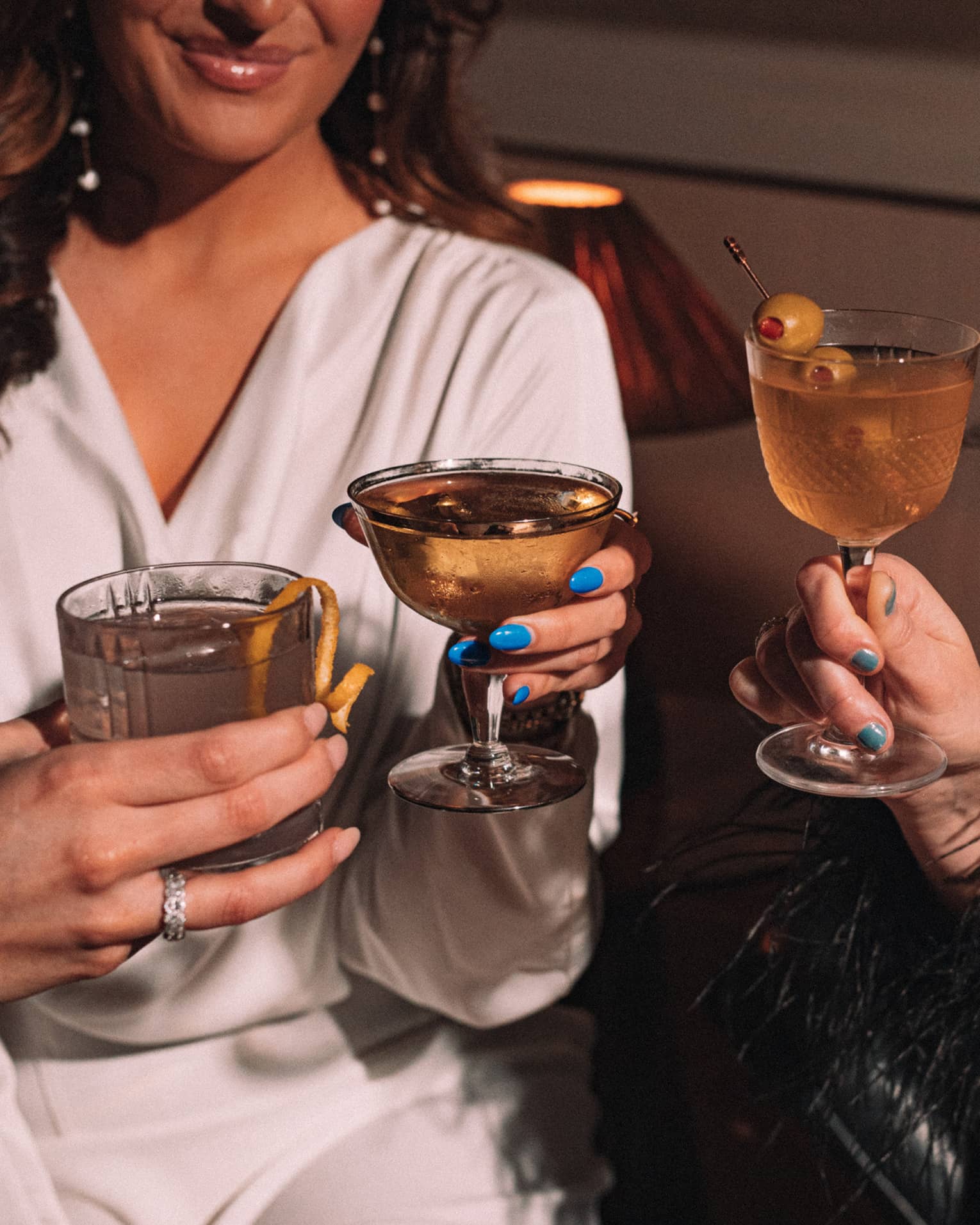 Three smiling guests share a toast with stemmed glasses and tumblers of cocktails with olive and lemon garnish.