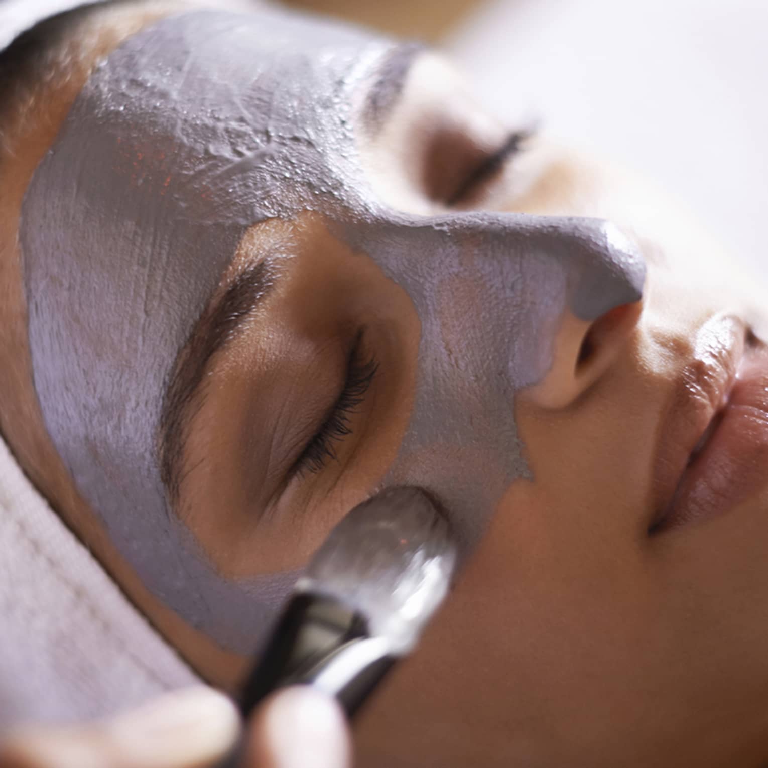 Woman with white towel around hair closes eyes as spa attendant applies grey face mask with paintbrush