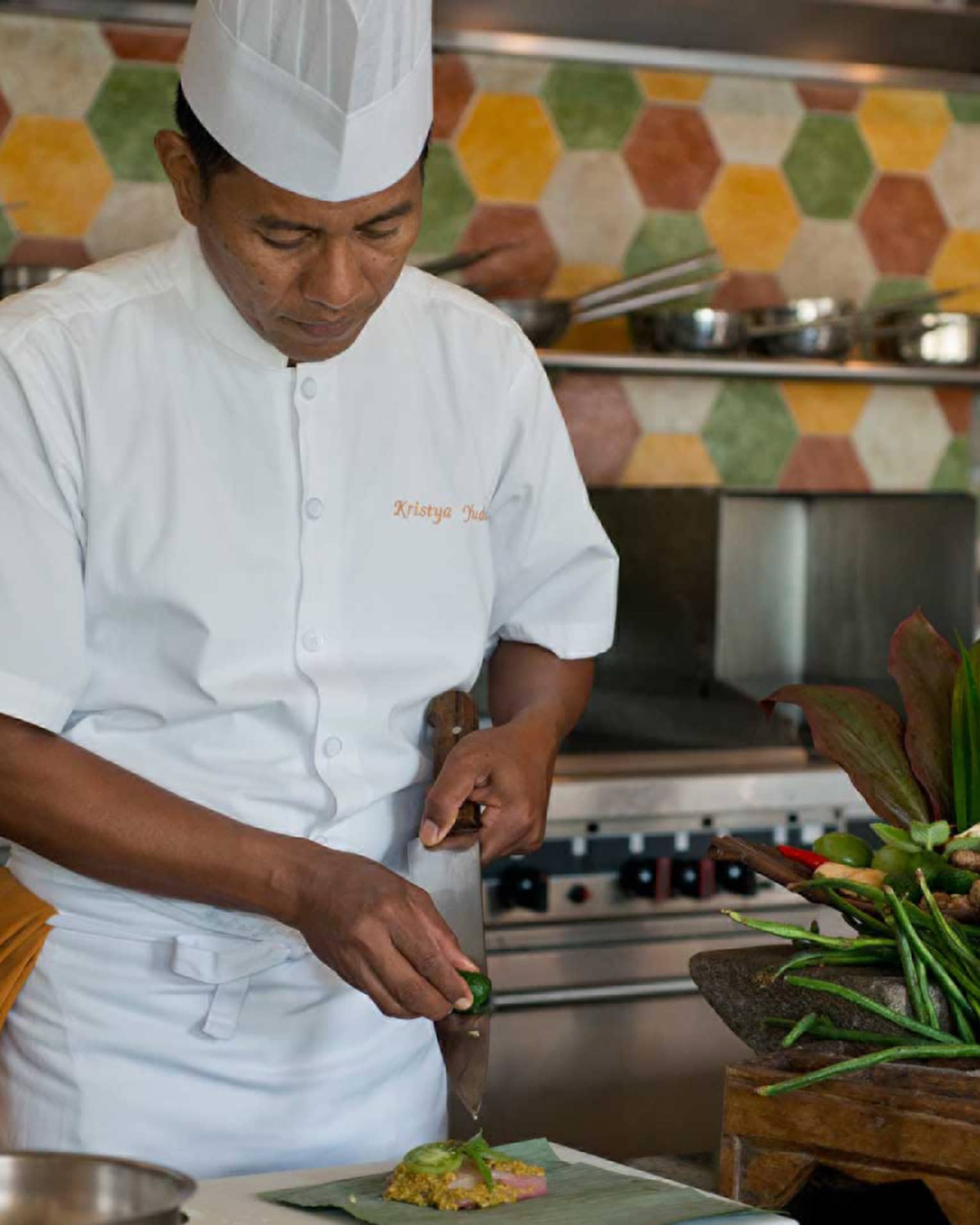 Chef in white uniform squeezes lime on large kitchen knife over dish, next to heads of fresh lettuce and whole peppers