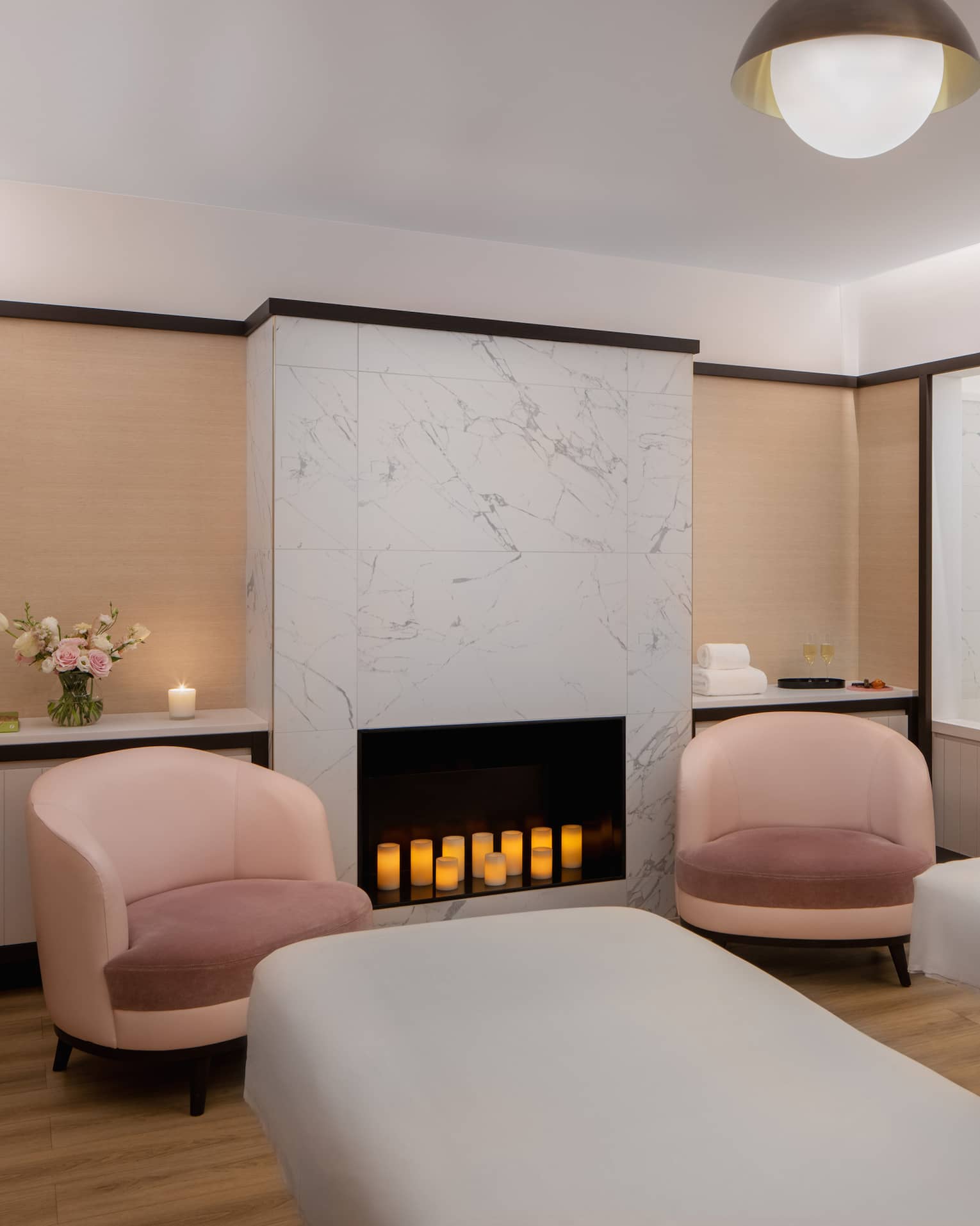 Couple's spa suite with a white-tiled fireplace filled with lit candles, an inset bathtub, two pink arm chairs, two side-by-side treatment tables and a pair of white robes