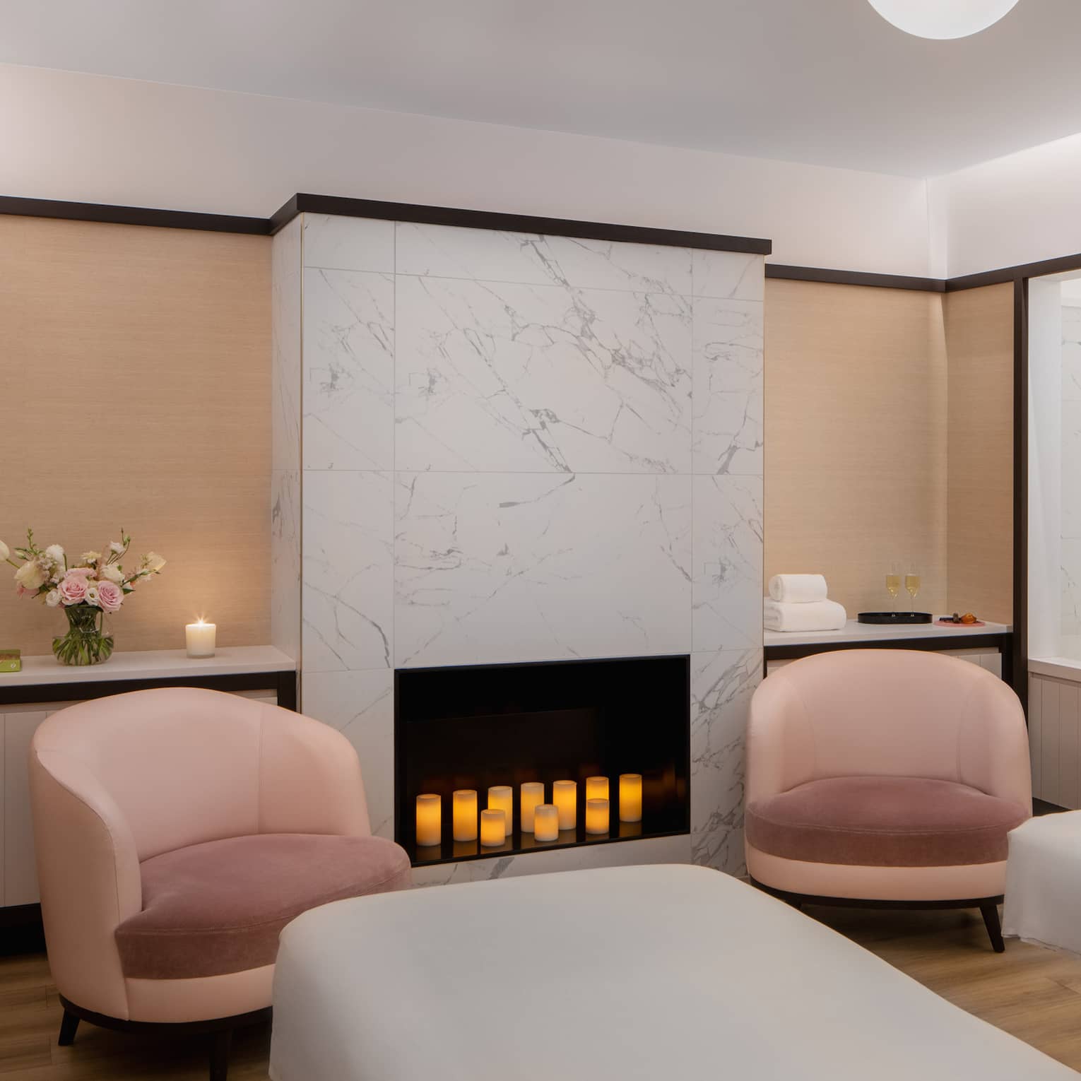 Couple's spa suite with a white-tiled fireplace filled with lit candles, an inset bathtub, two pink arm chairs, two side-by-side treatment tables and a pair of white robes