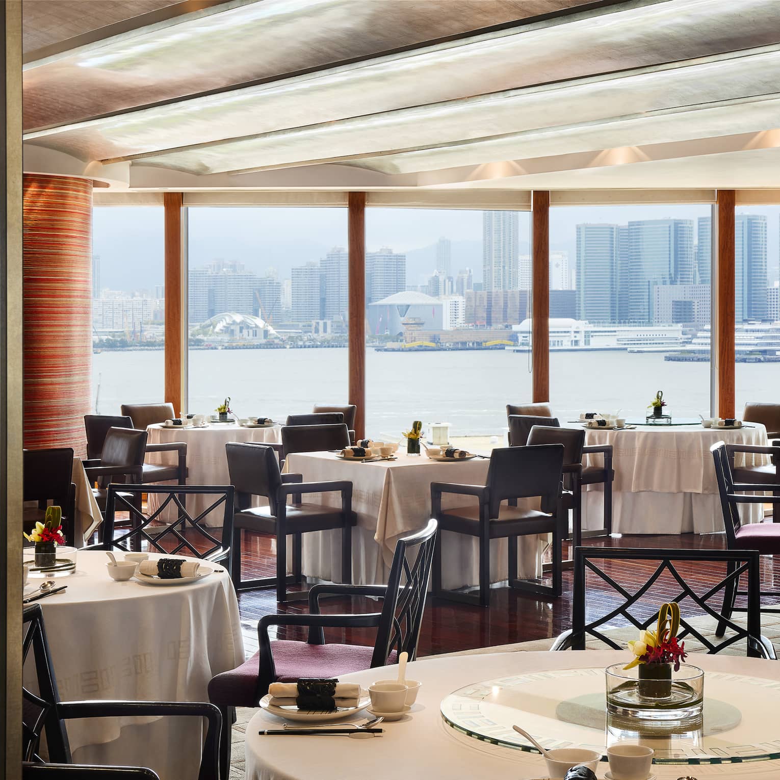 An elegant dining room with tables set with flowers and place settings, overlooking the city from across a body of water.