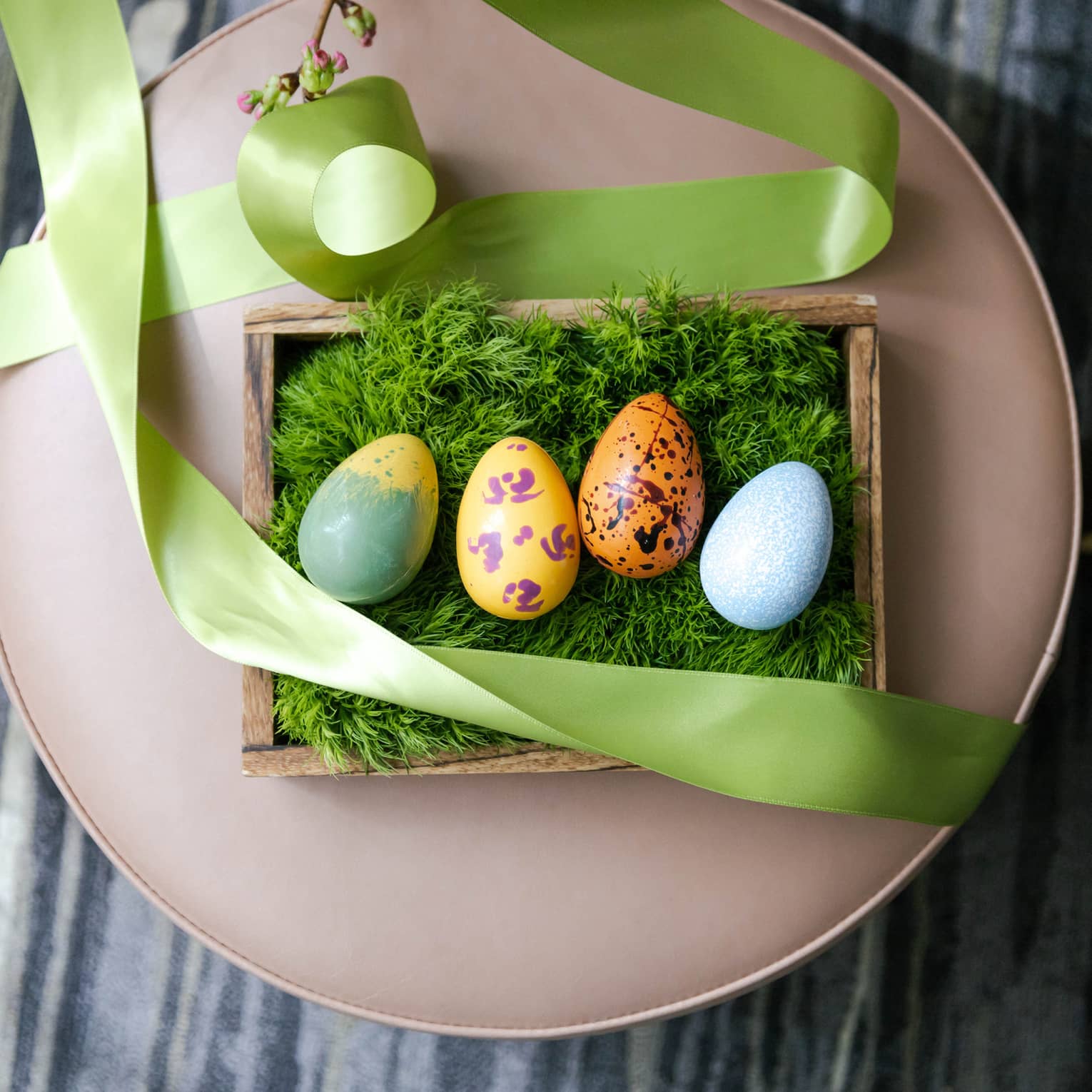 Four multi-colored chocolate Easter eggs on a bed of green moss set in a wooden box with a light-green ribbon draped around it