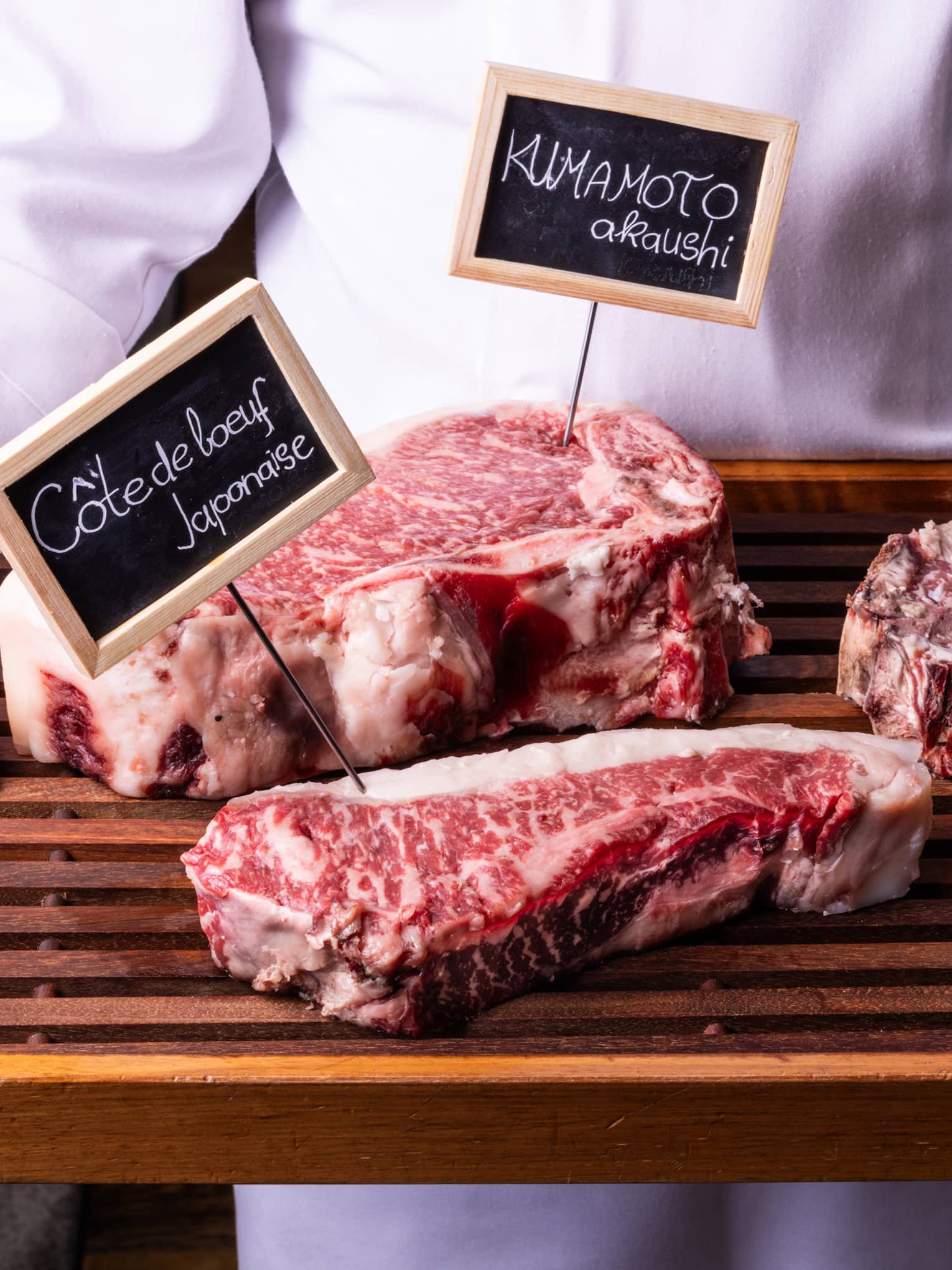 Close-up view of a wood tray with three labelled cuts of well-marbled beef, held by person in white chef's coat.