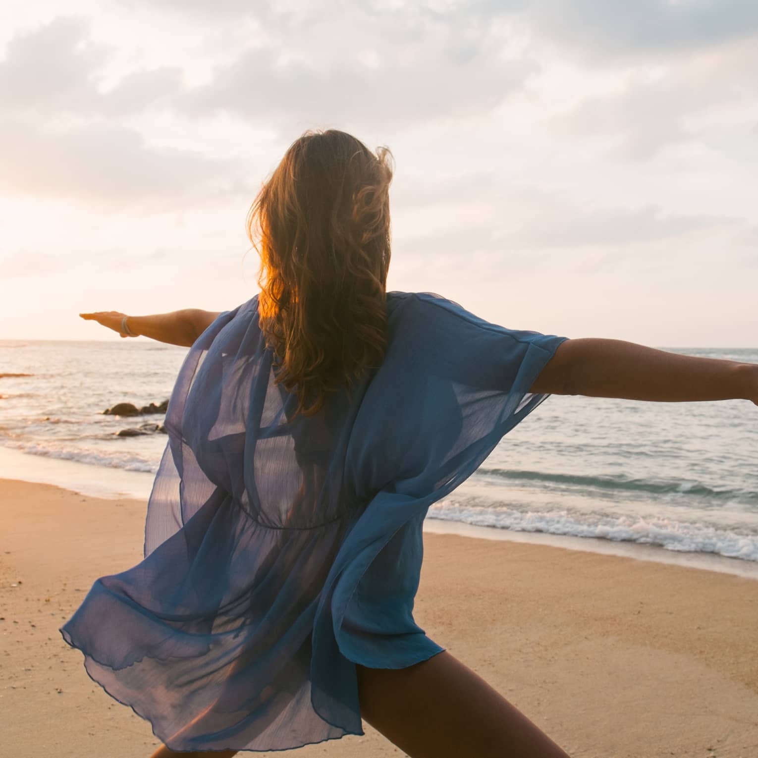 Woman wearing sheer beach cover kneels with arms outstretched in yoga pose on sandy beach by water