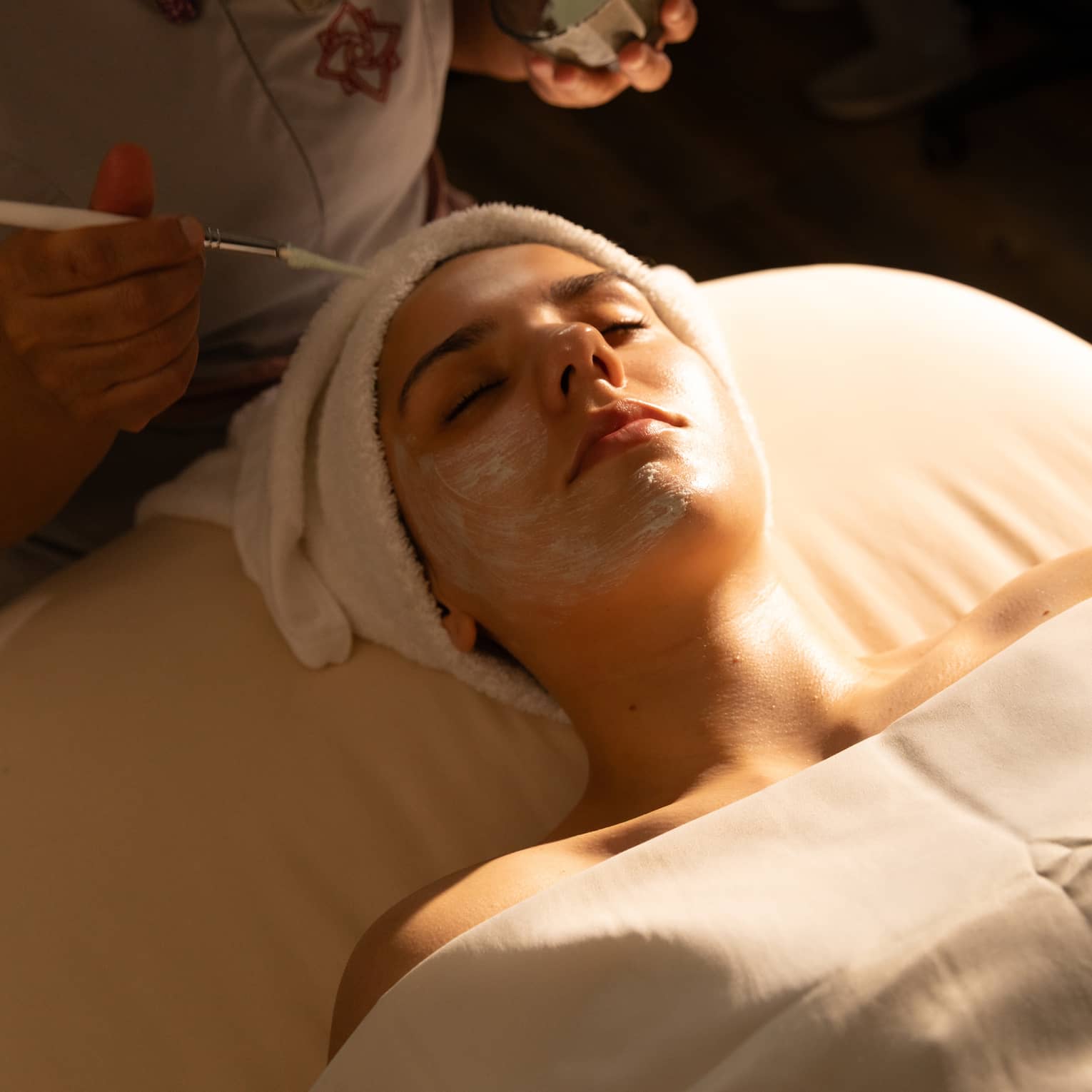 A person with draping over their body and a towel wrap on their head lies on a spa treatment table and receives a facial treatment.