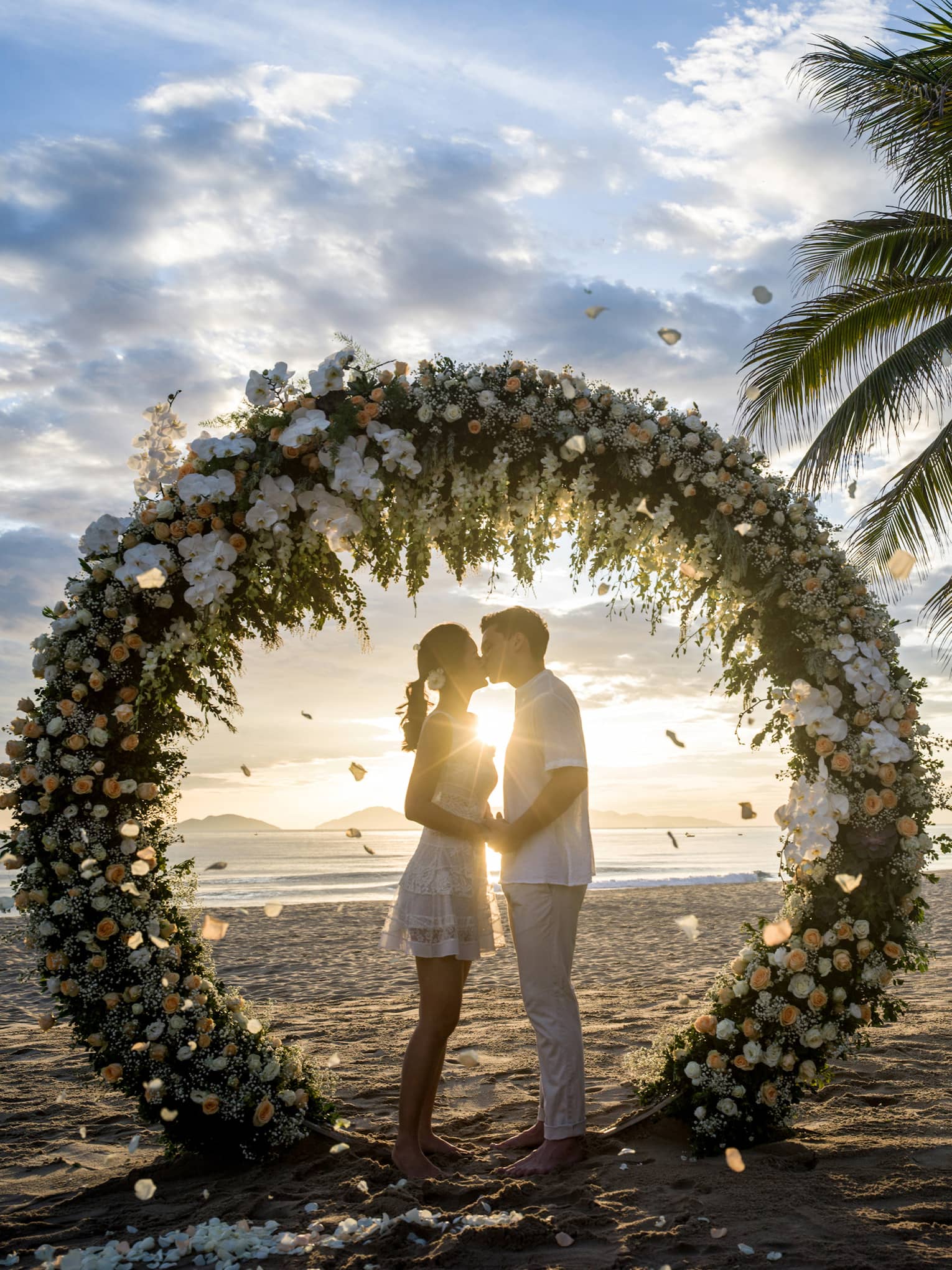 Bride and groom kiss under round wedding altar with flowers on beach at sunset
