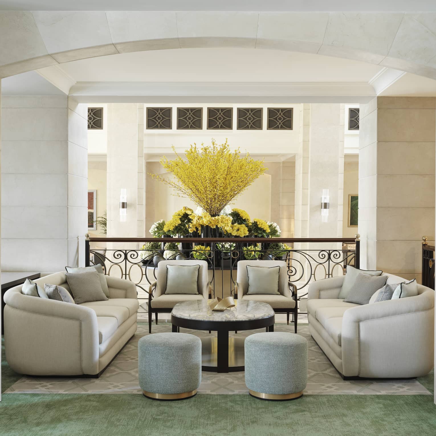 Two white sofas, two white arm chairs, two light-blue upholstered ottomans and an oval marble-top coffee table fill the center of a lounge area within a hotel lobby