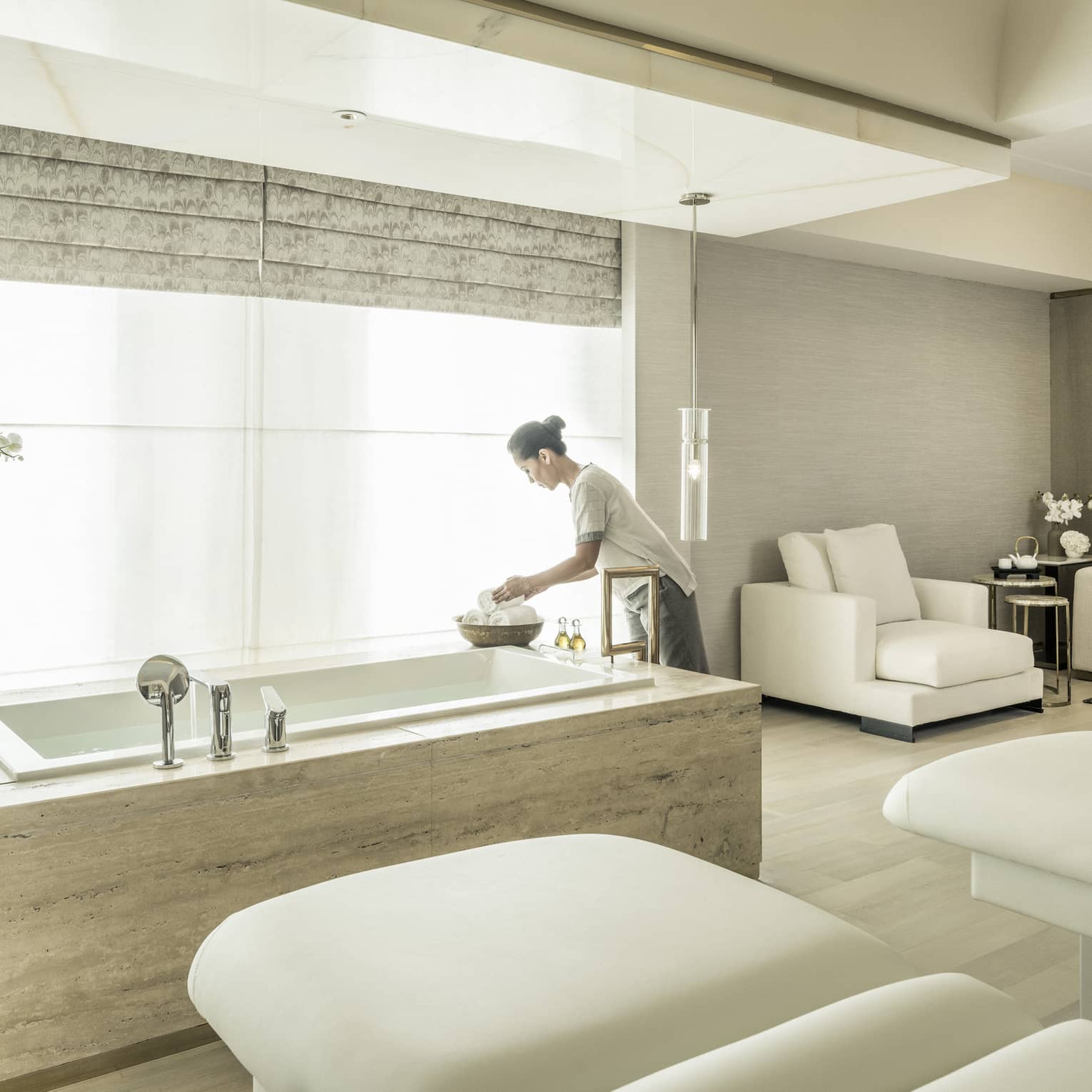 A spa attendant arranges some products in the spa suite, with two massage beds and a soaking tub in front