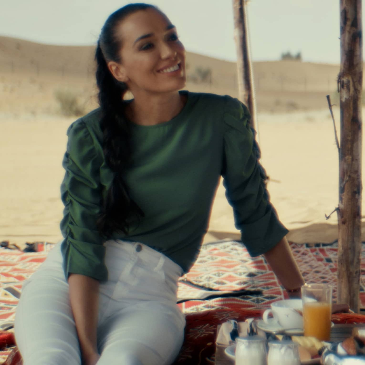 Two smiling guests lounge on a shaded blanket in the desert as a third person bends over a tray of drinks and food.