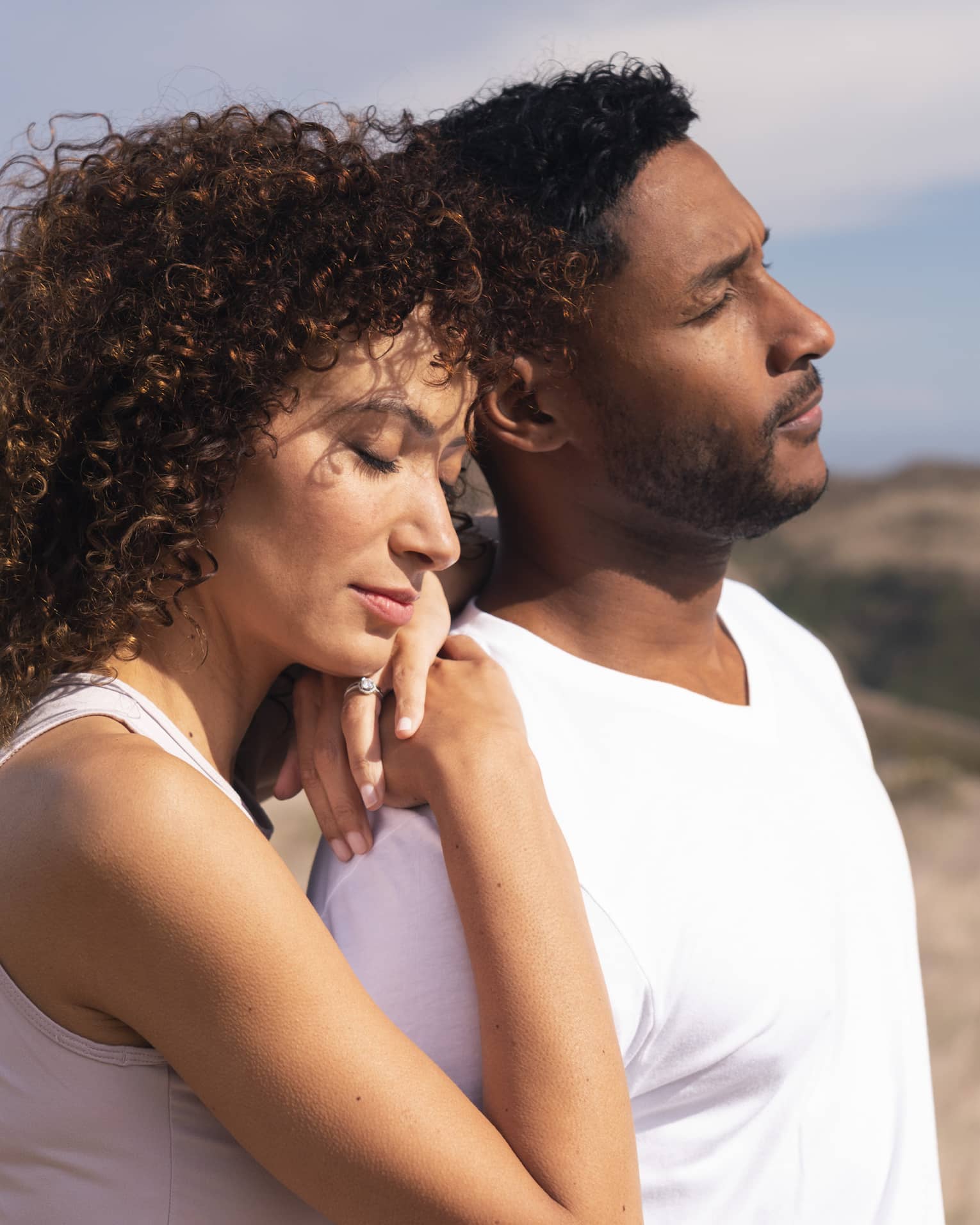 Two people both wearing white t-shirts stand together in a field with their eyes closed, one with their arms on the other's shoulders
