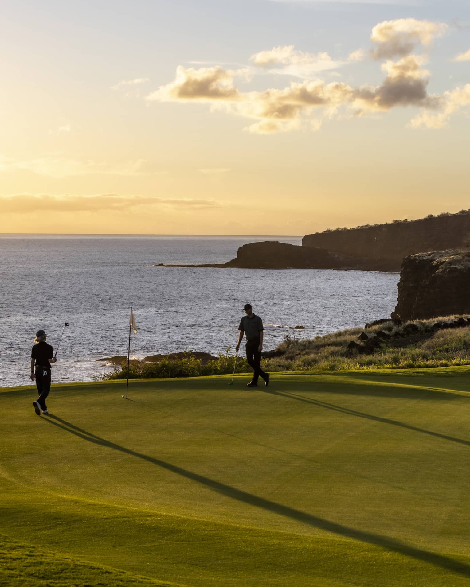The setting sun silhouettes a pair of golfers enjoying a game of golf. The golf course is on top of a cliff beside the ocean.