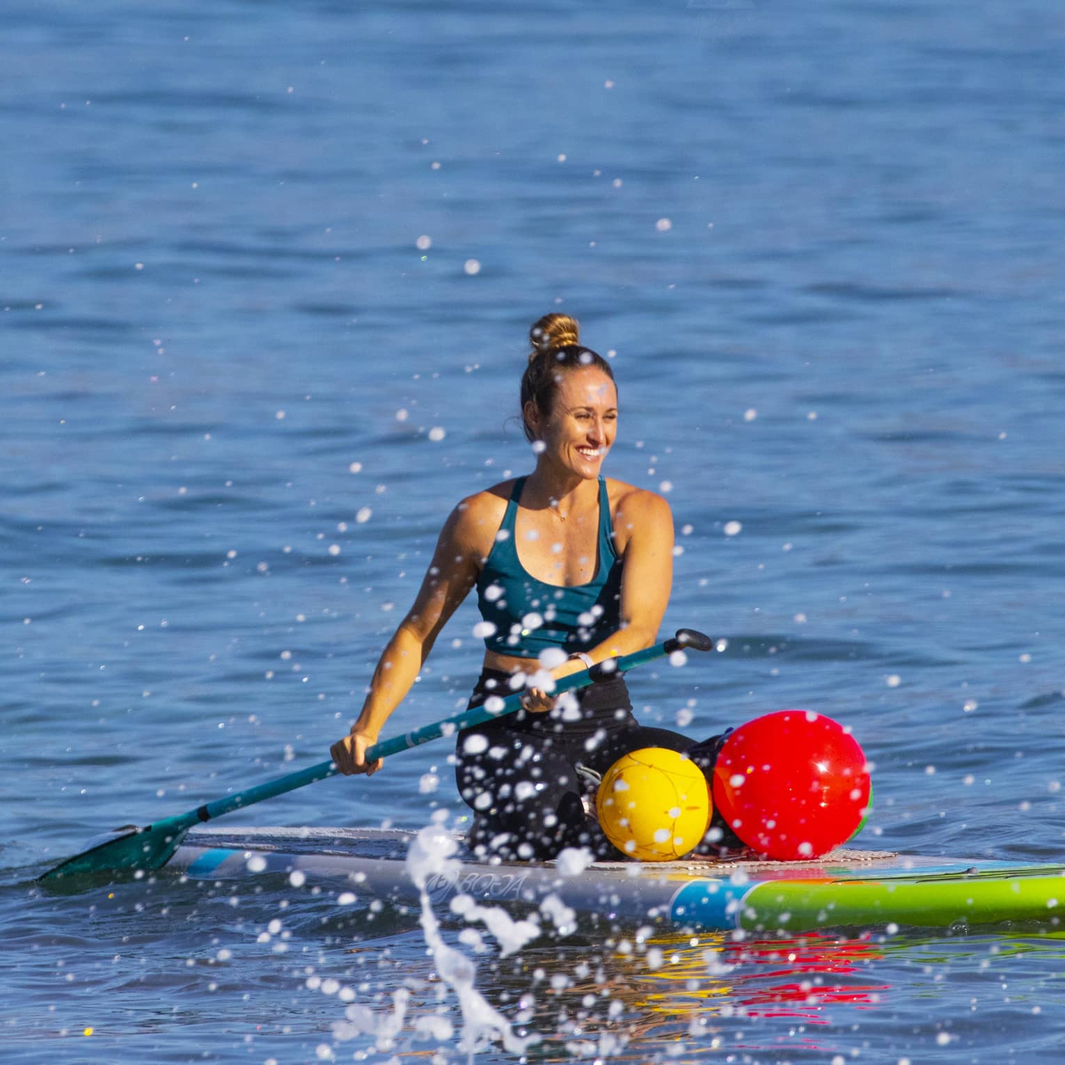 Close-up front view of a smiling guest kneeling on a stand-up paddleboard on calm water, a frothy wake in the foreground.
