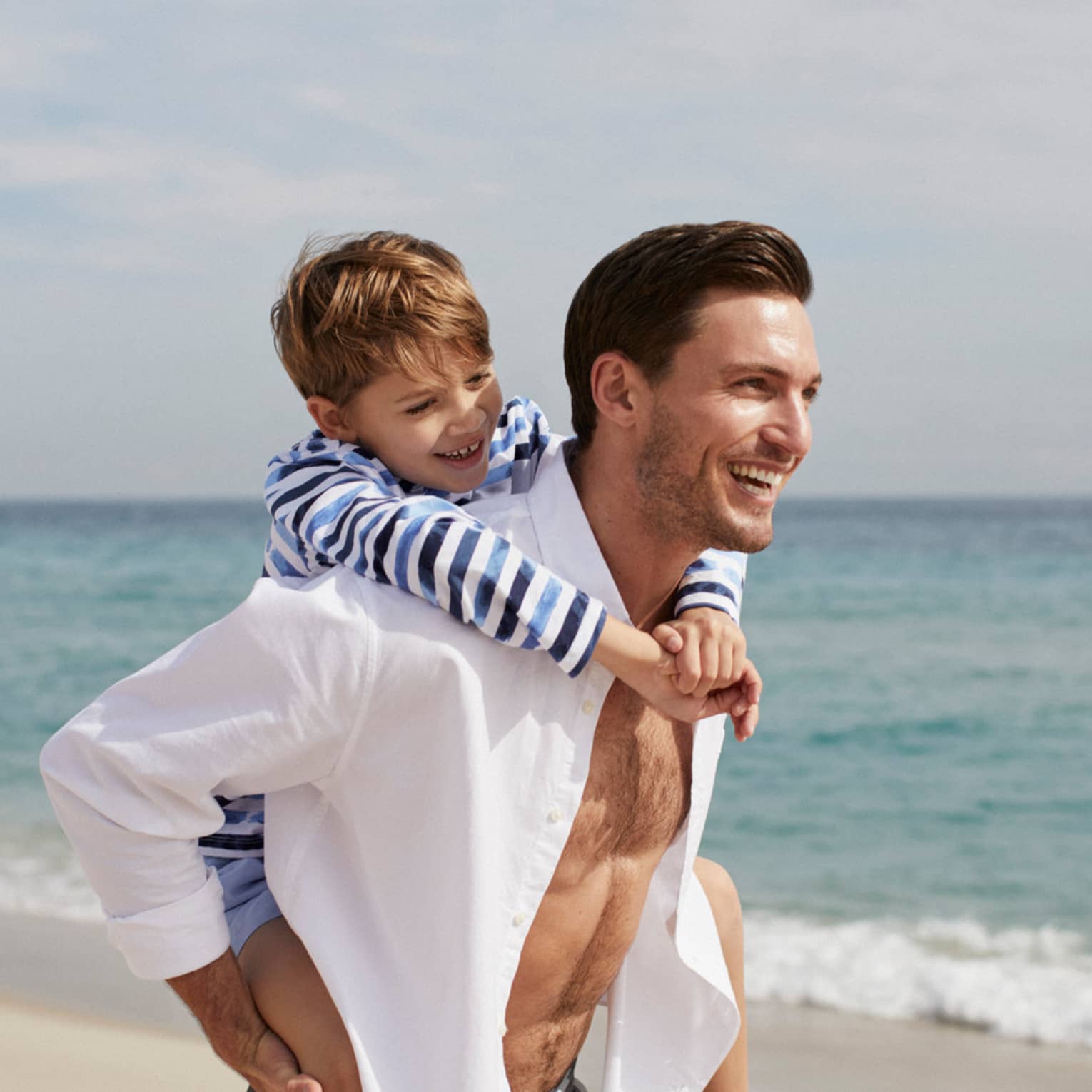 A man smiles as he carries a young boy on his back while walking on a beach
