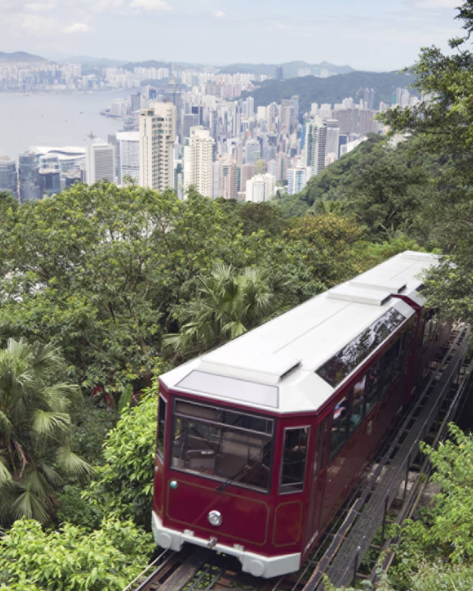 The red Peak Tram ascends a lush, green hillside with a sprawling cityscape in the background. Tall skyscrapers rise into the skyline under a cloudy sky.