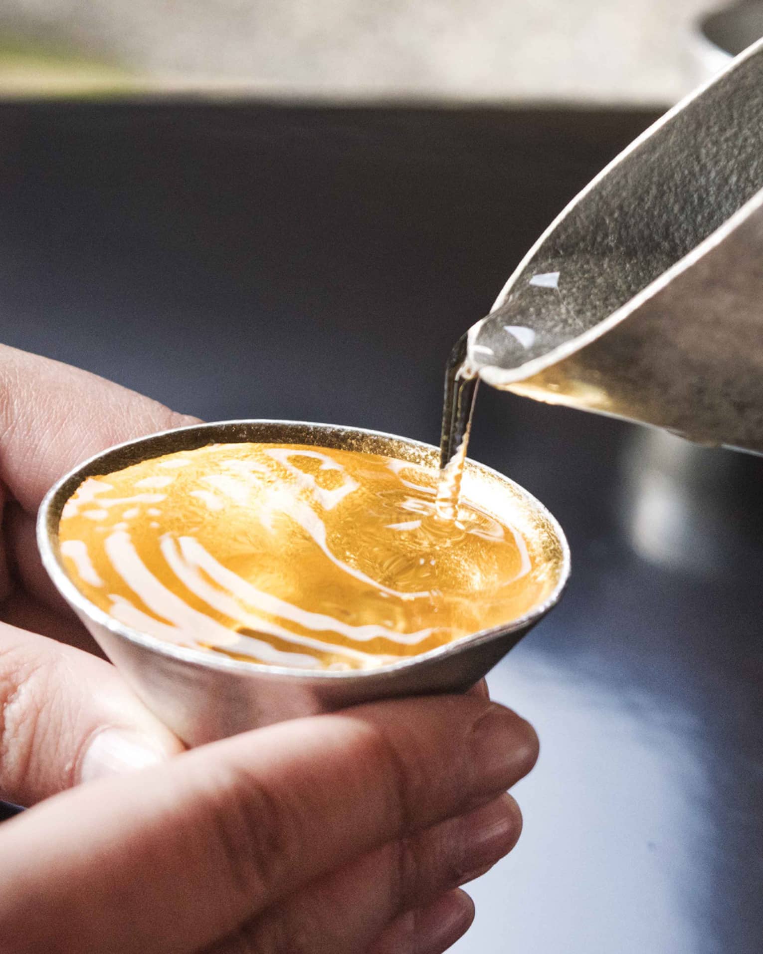 Clear liquid taking on amber colour as it is poured from a hammered metal spout into small cone-shaped cup held by two hands