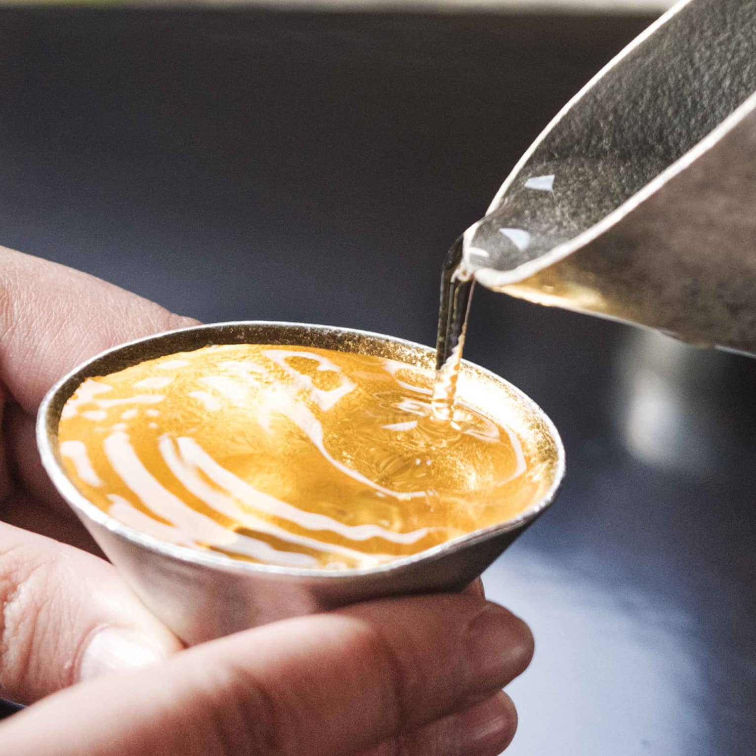 Clear liquid taking on amber colour as it is poured from a hammered metal spout into small cone-shaped cup held by two hands