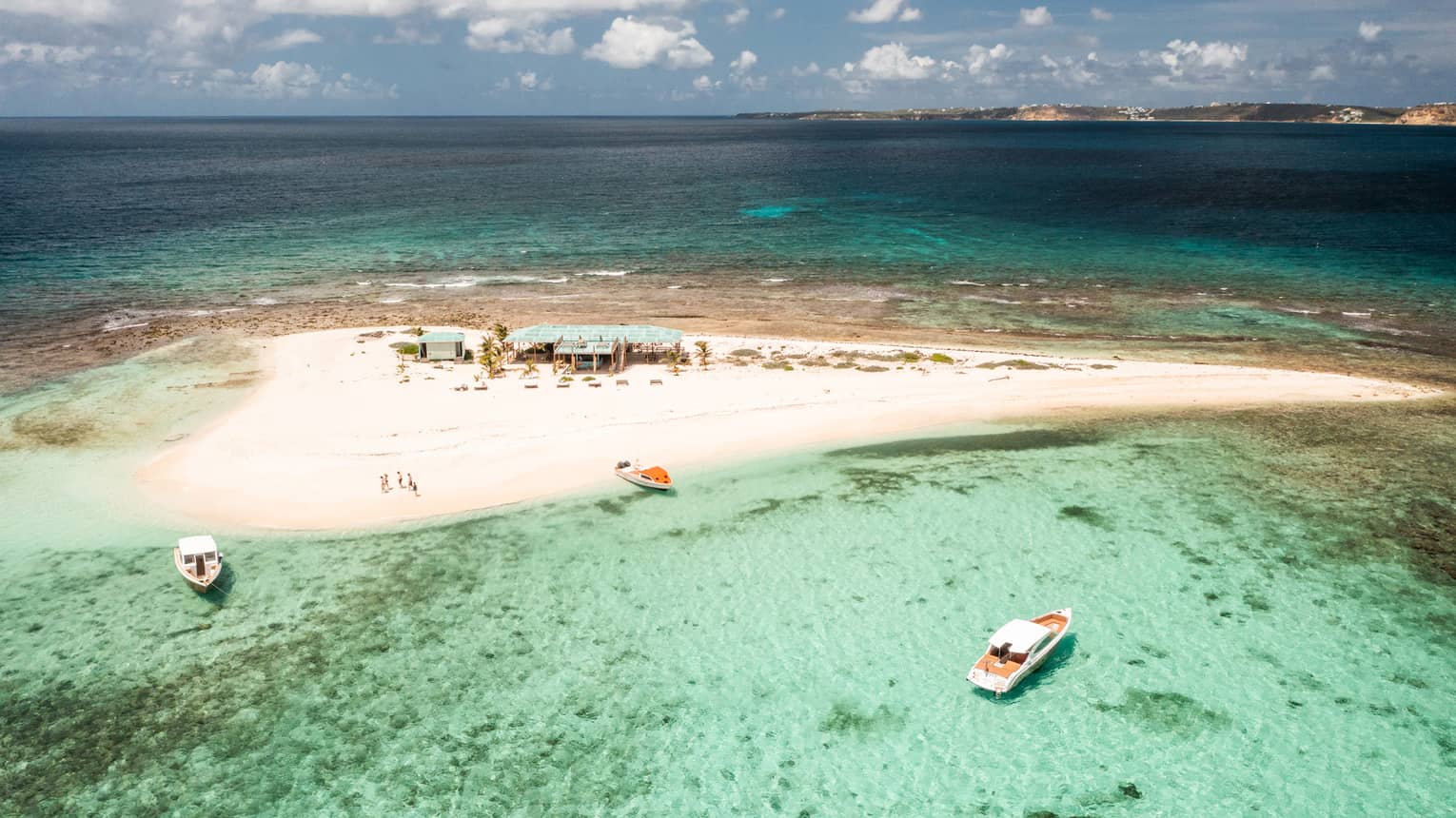 A small sandy island surrounded by crystal clear water and a few boats