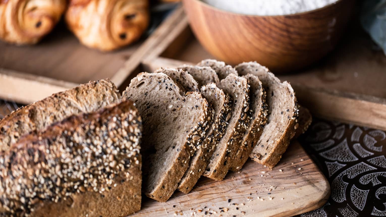 Loaf of rye bread sliced half sliced half whole with a bowl of flour and a couple croissants in the background