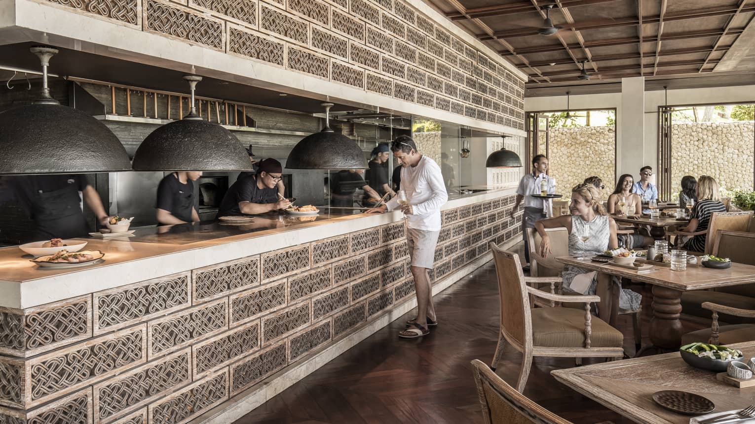 Restaurant guests watch as chefs plate entrees on ledge of open kitchen under accent wall